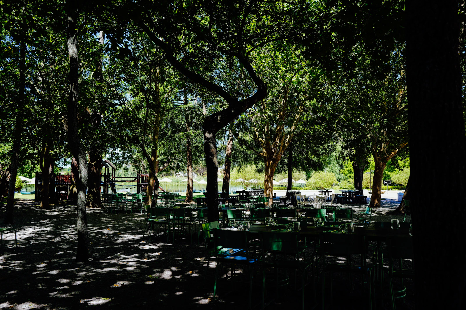 Green tables and chairs sit beneath deep shade beside water.