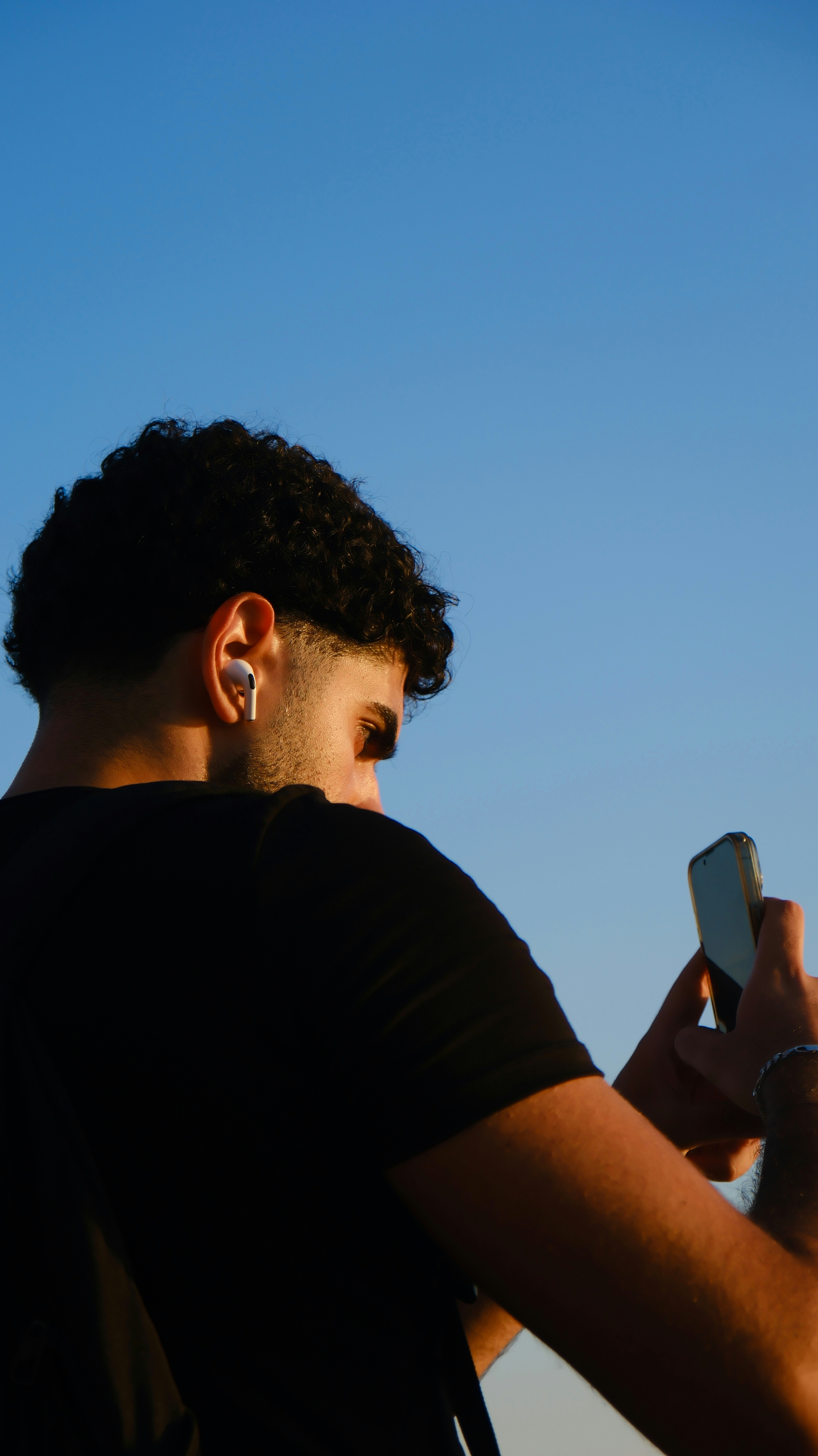 Uomo con capelli ricci che indossa gli auricolari e tiene il telefono in mano.