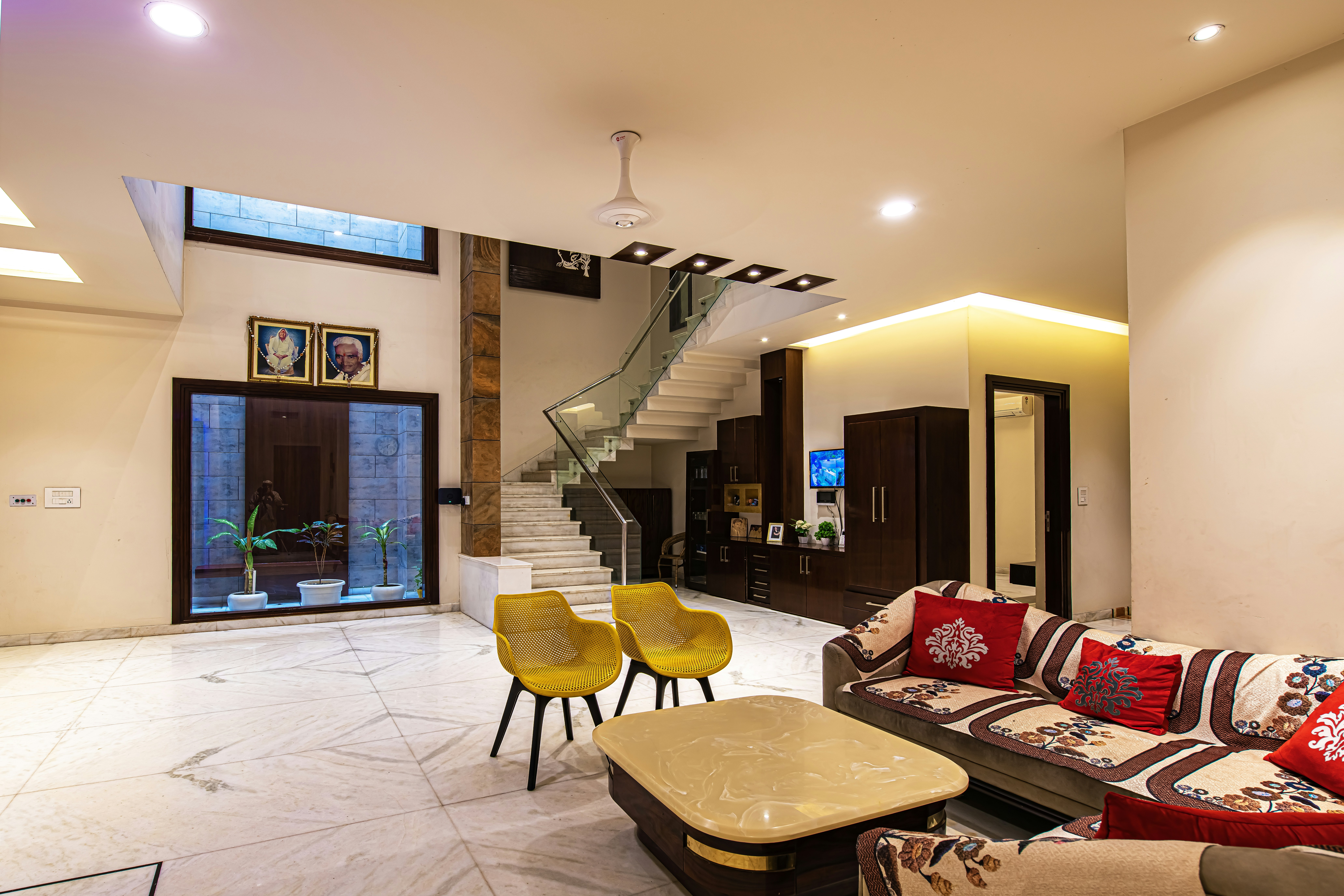 A spacious, modern multi-level living room features a grand marble staircase, warm lighting, dark wood cabinetry, yellow chairs, and a patterned sofa.