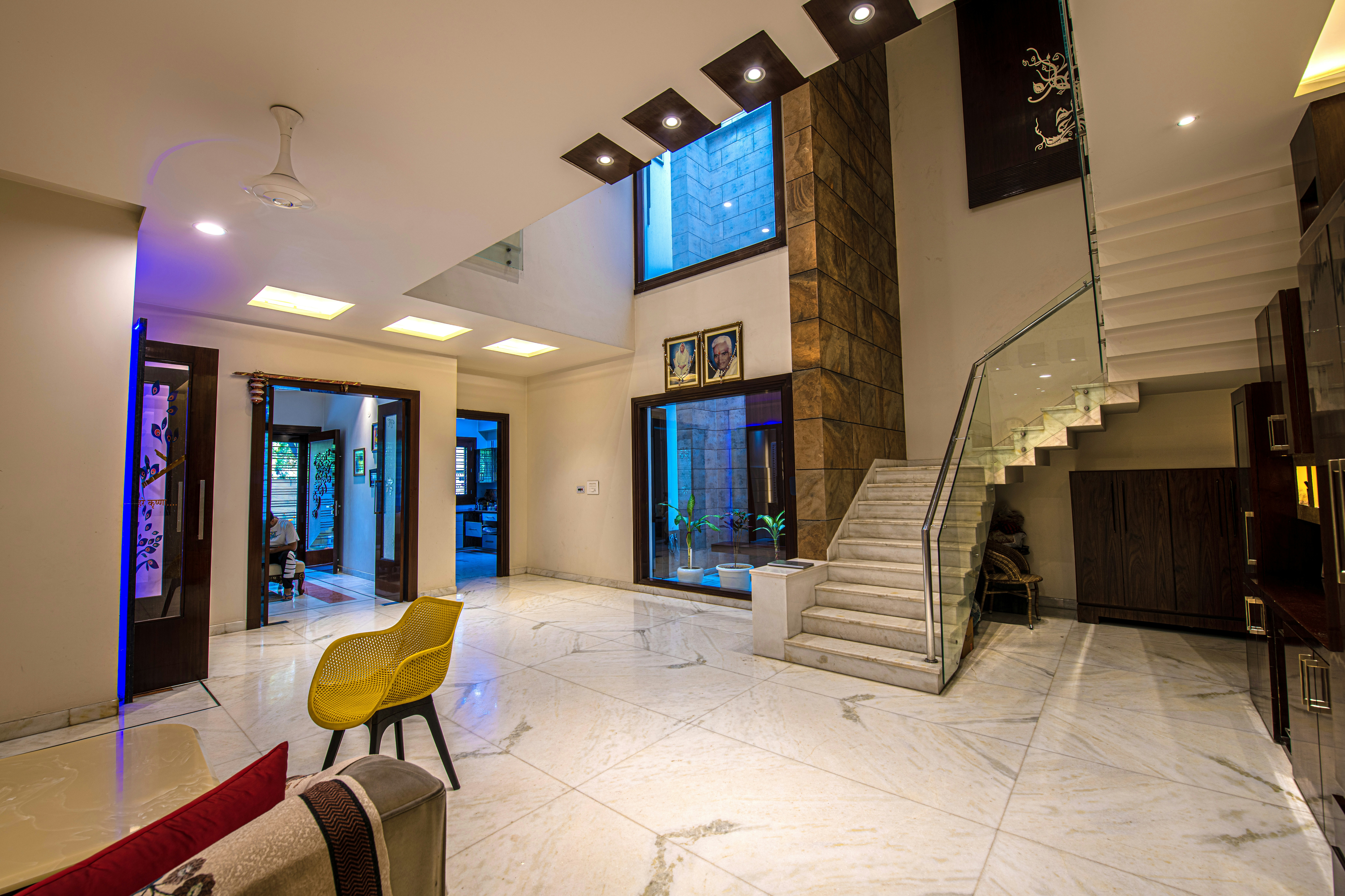 A spacious, modern double-height entrance hall with white marble floors, a grand staircase, stone wall accents, and blue mood lighting.