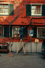 Man painting a red building while standing on a ladder.