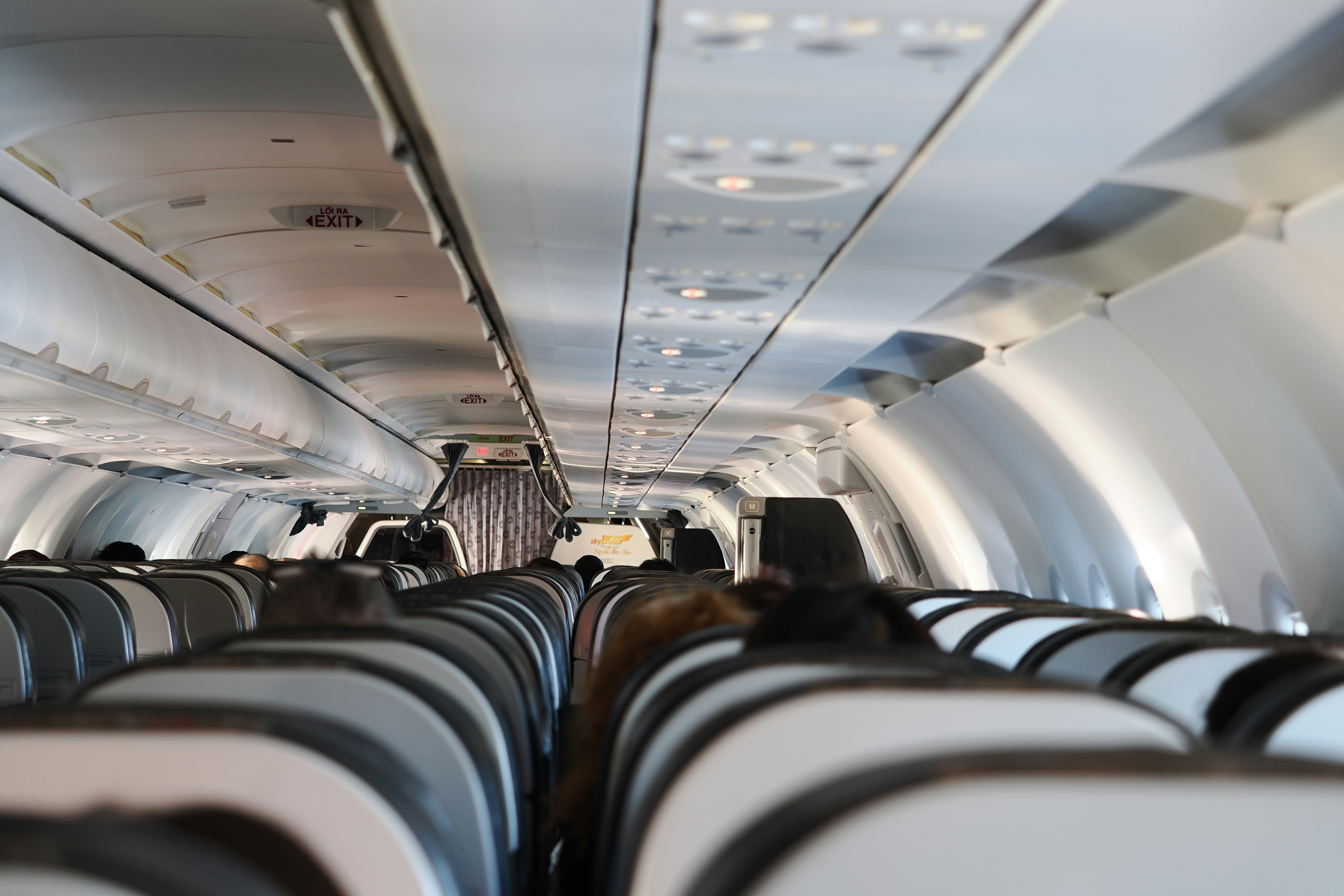 A view from inside an airplane cabin, showing passengers looking concerned or anxious. Cabin crew members are visible in the aisle, talking to each other or to a passenger. The atmosphere suggests a disruption.