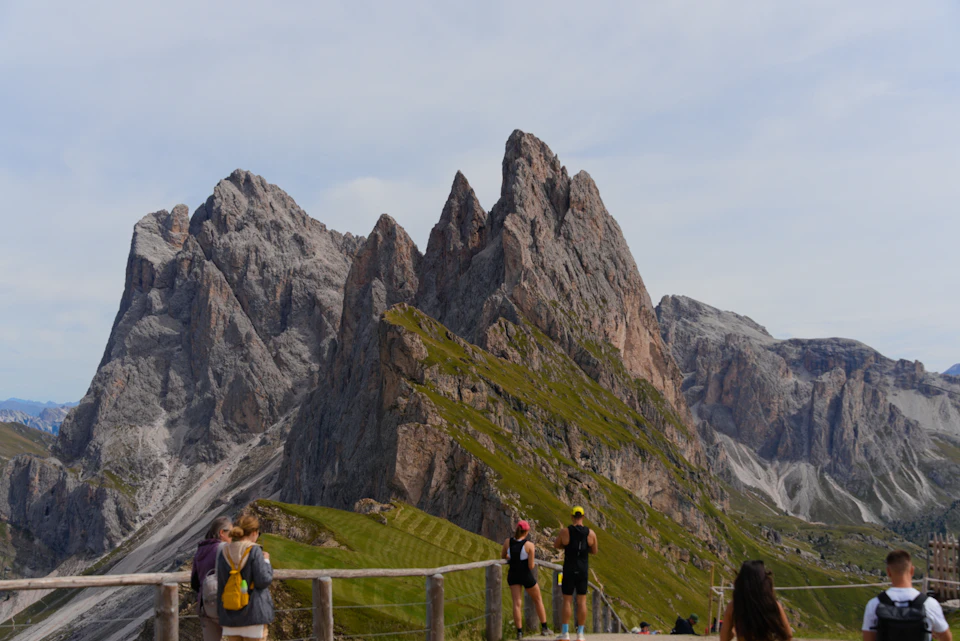 The image depicts a group of people standing on a wooden platform, overlooking a breathtaking mountain range with jagged peaks and valleys. The mountains are covered in lush green vegetation, contrasting with the clear blue sky above. Some individuals in the group are wearing backpacks, suggesting they may be hikers or adventurers exploring the area.