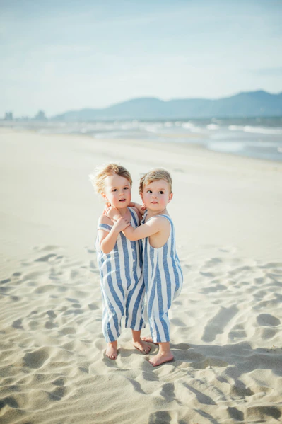 Two young children in striped overalls on a sandy beach.