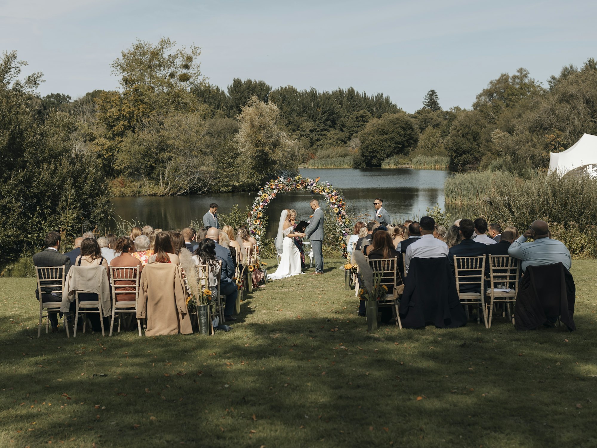 Outdoor summer wedding ceremony beside a lake with guests seated under sunny skies