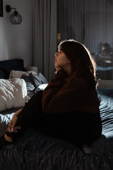 A young woman sits on a bed in dim lighting.