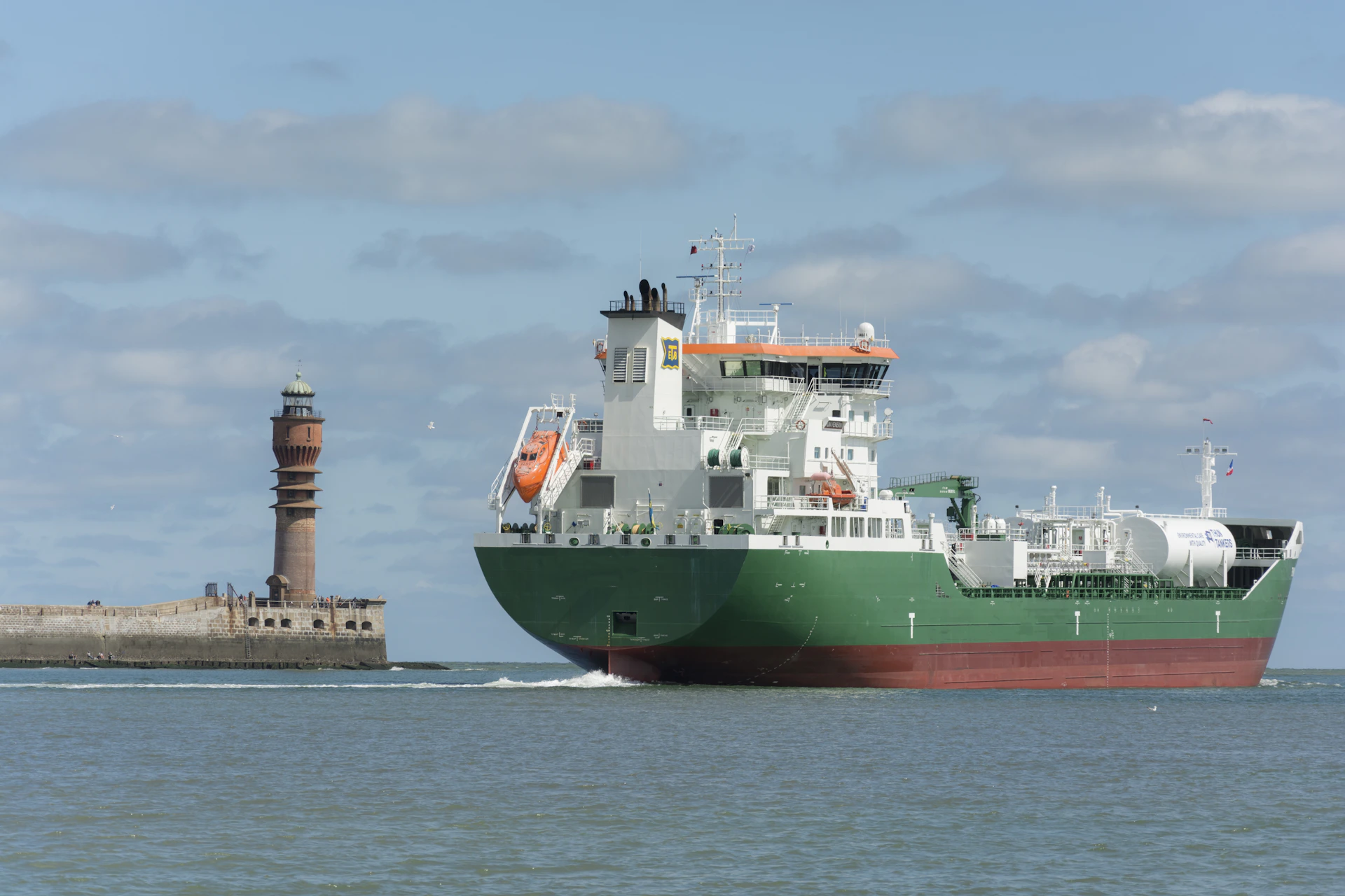 A large green ship sails past a lighthouse