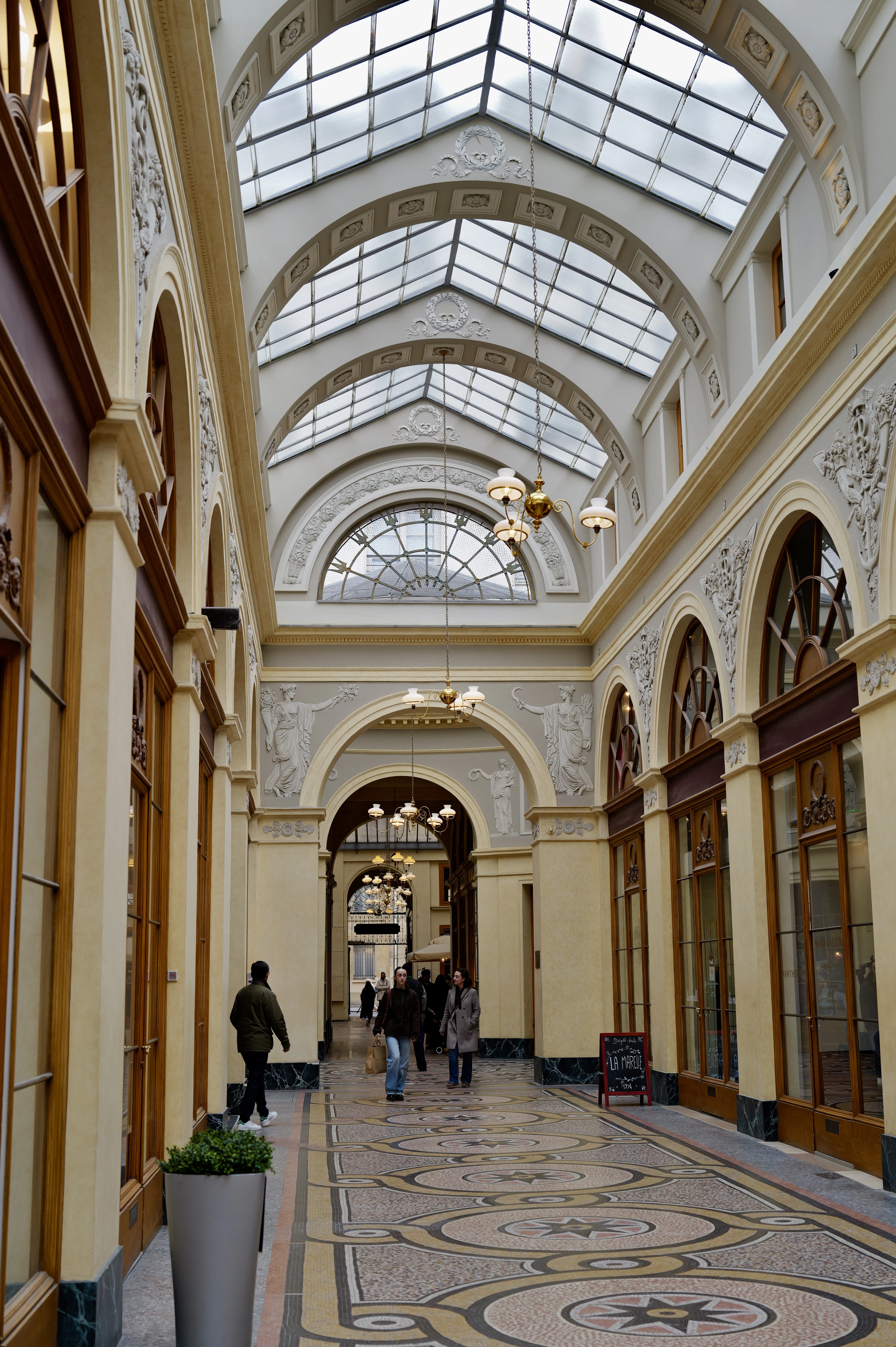 The interior of the Galerie Vivienne in Paris, a nineteenth-century covered passage lit through an arched glass roof, its mosaic floor patterned with star medallions and its pale walls set with sculpted classical figures and brass lanterns above the shopfronts