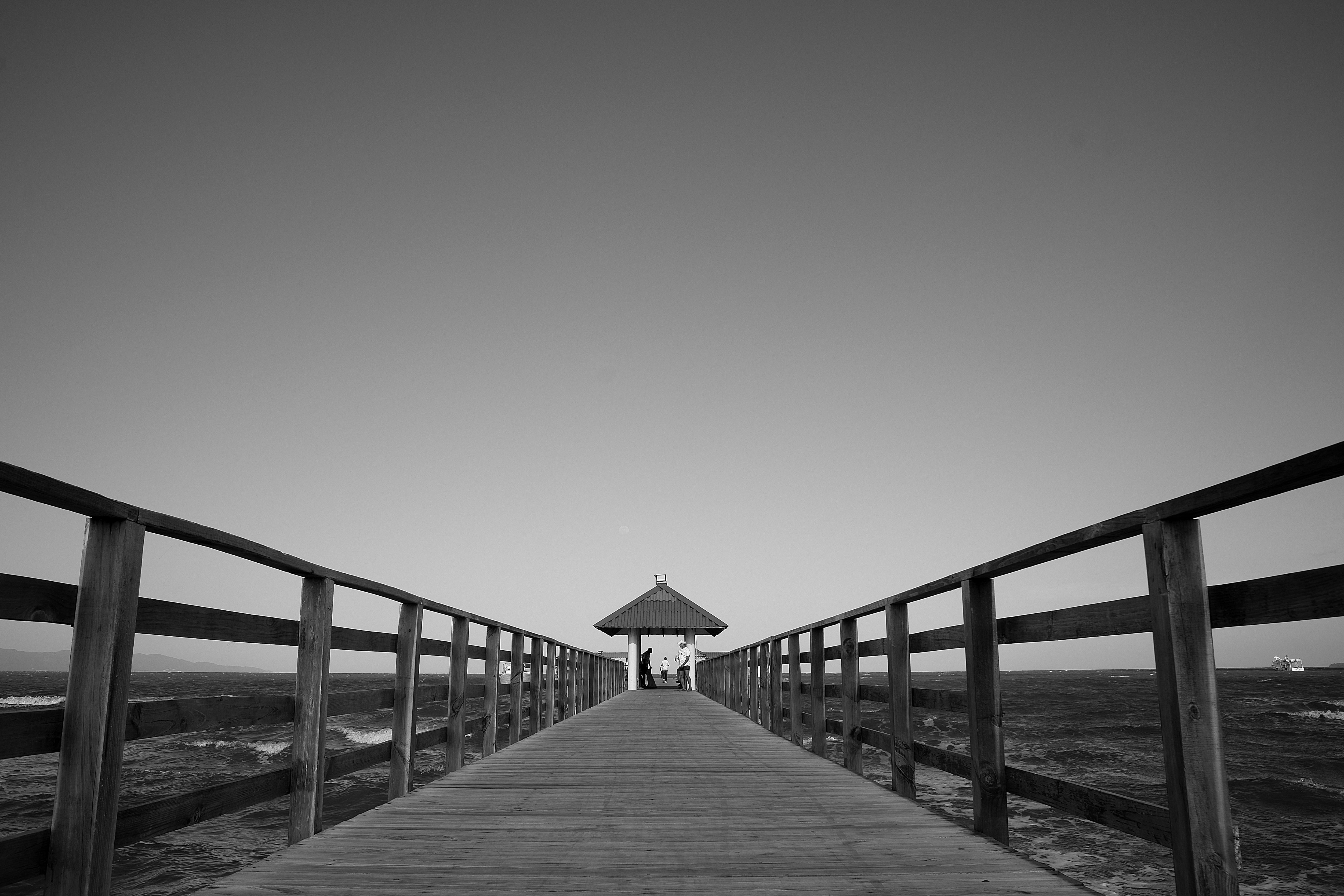 A black-and-white pier stretches toward a distant shelter, centering a calm, vanishing point over calm waters.