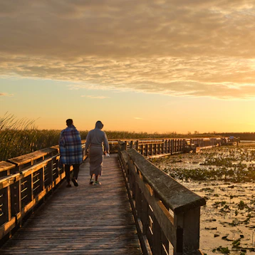 Two people walking with Jesus on a wooden boardwalk at sunset.