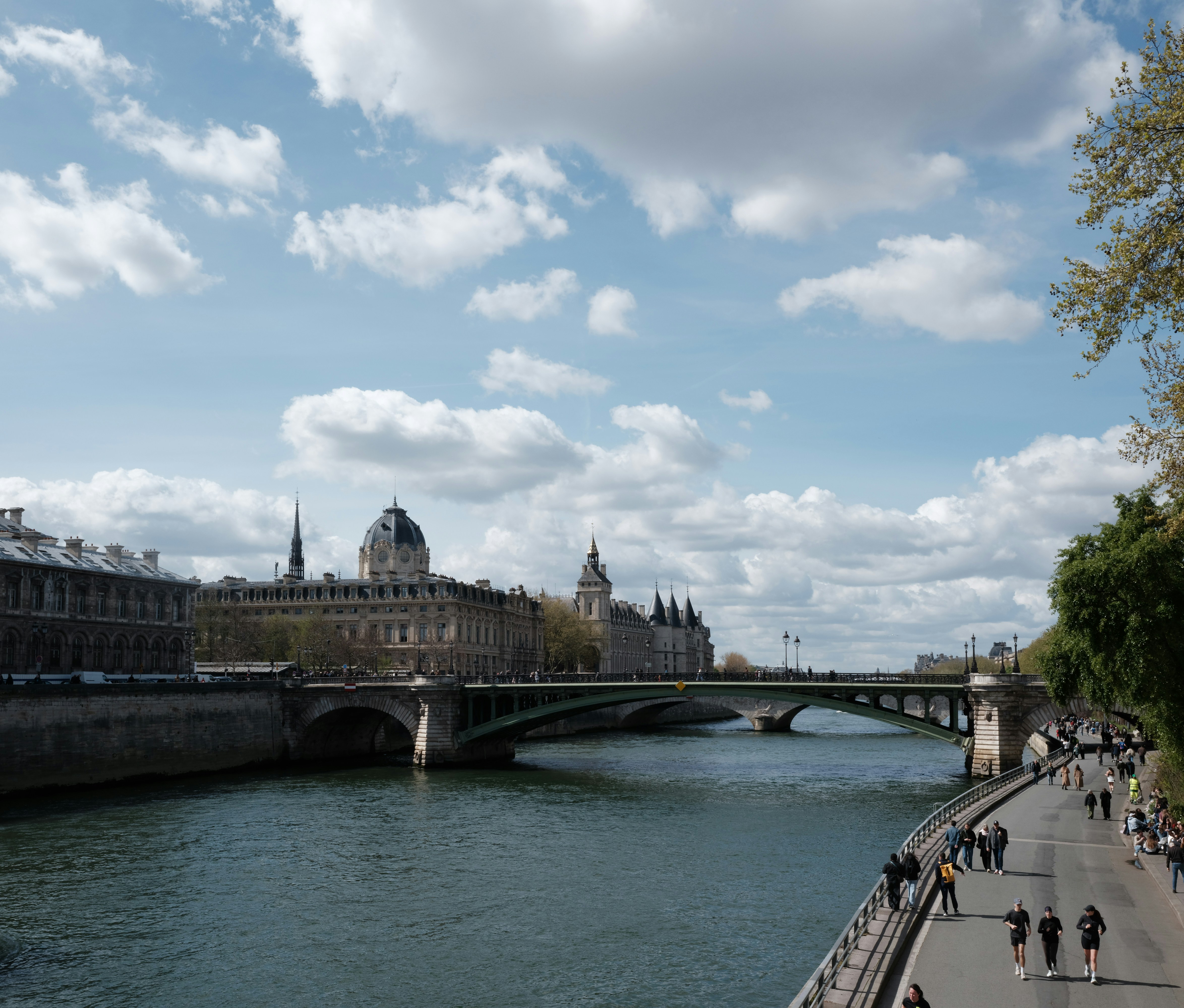 River flowing by buildings with people walking on path.