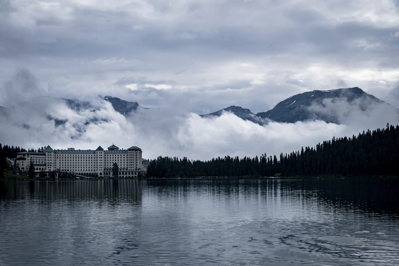 A grand mountain chateau hotel sitting at the edge of a still glacial lake with forested mountains and low cloud behind, the building's facade reflected in the water