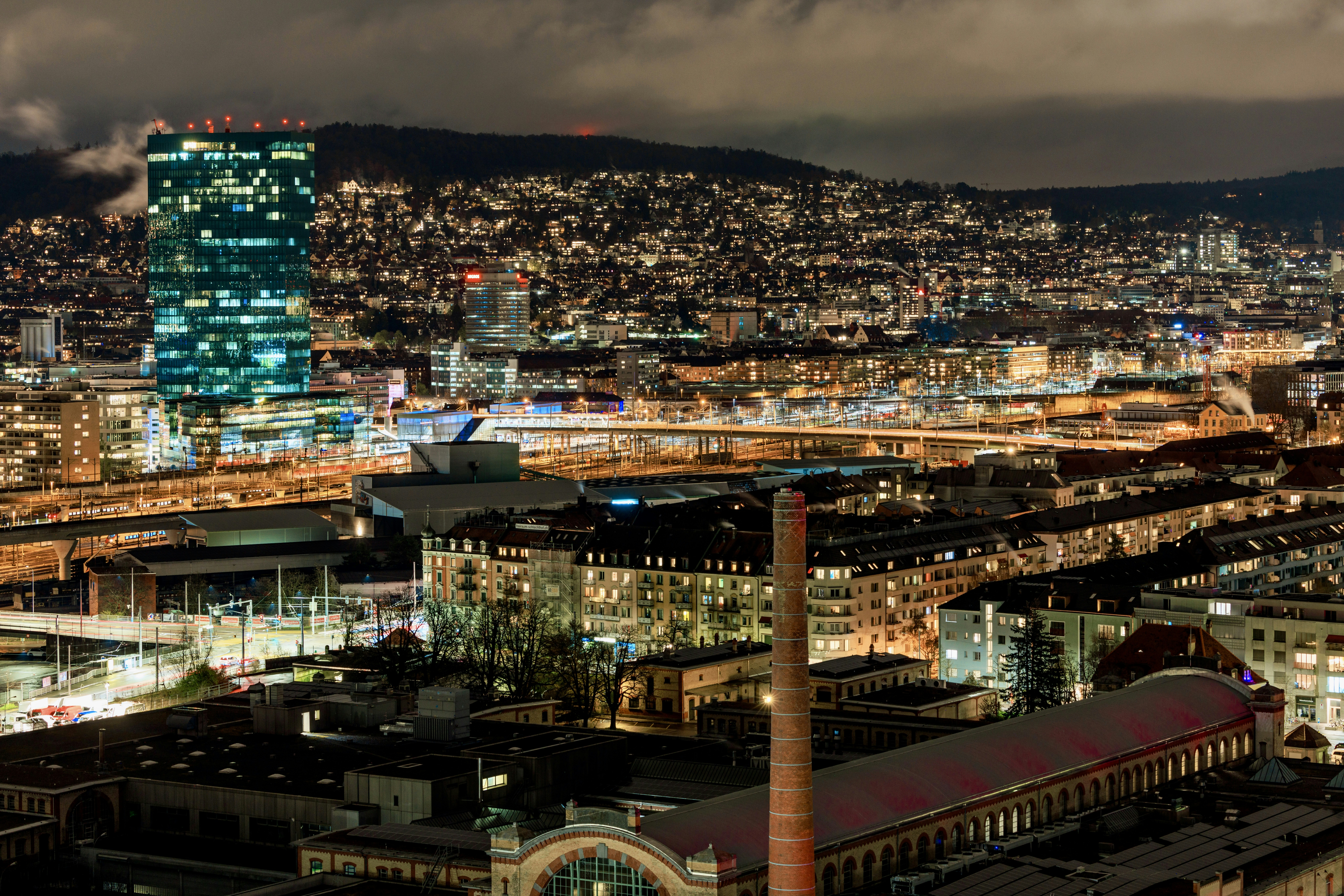 Paesaggio urbano di notte con edifici illuminati e colline.