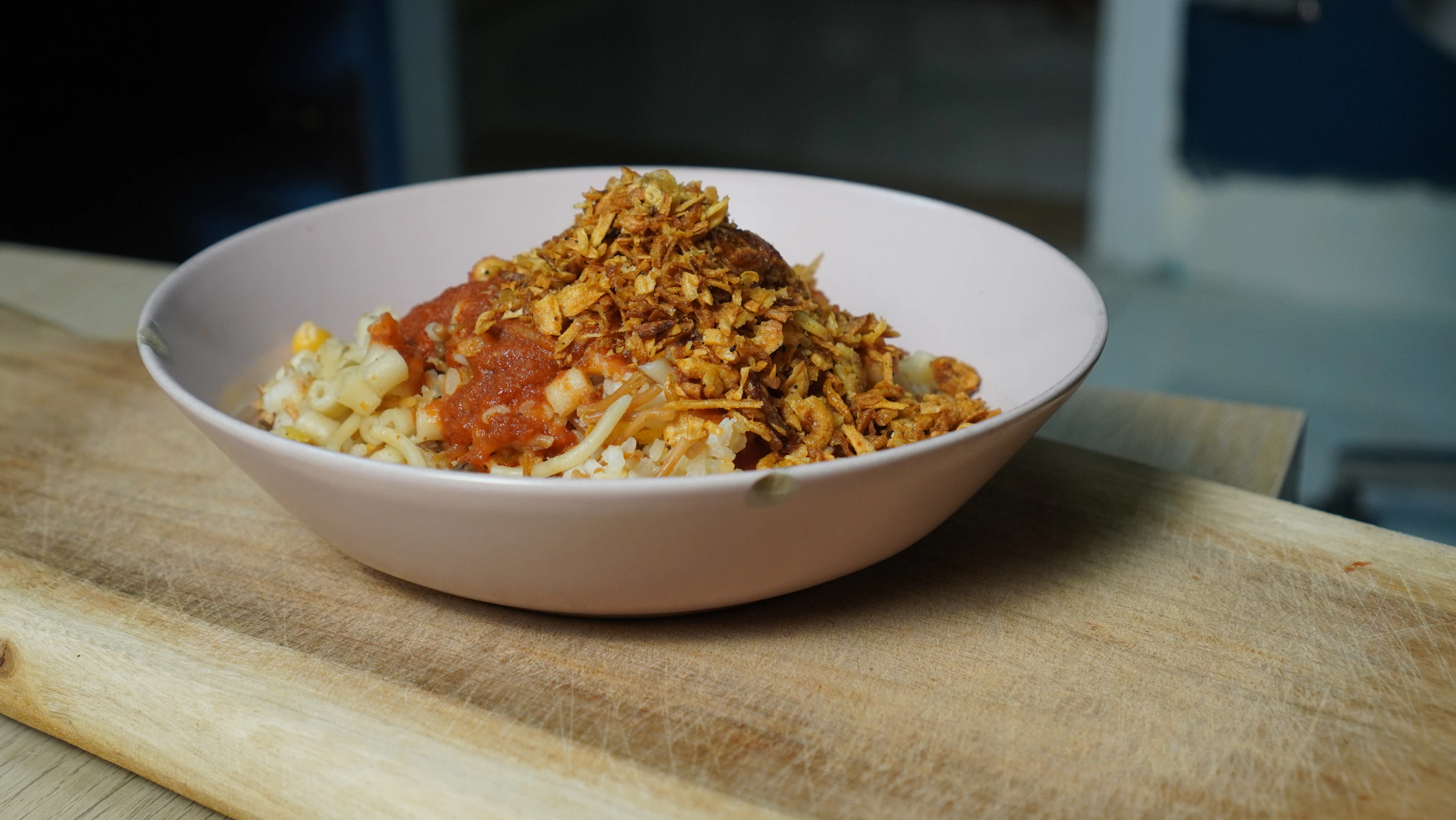 A bowl of Egyptian koshari — macaroni, rice and lentils under tomato sauce and a heap of crisp fried onions — set on a wooden board.