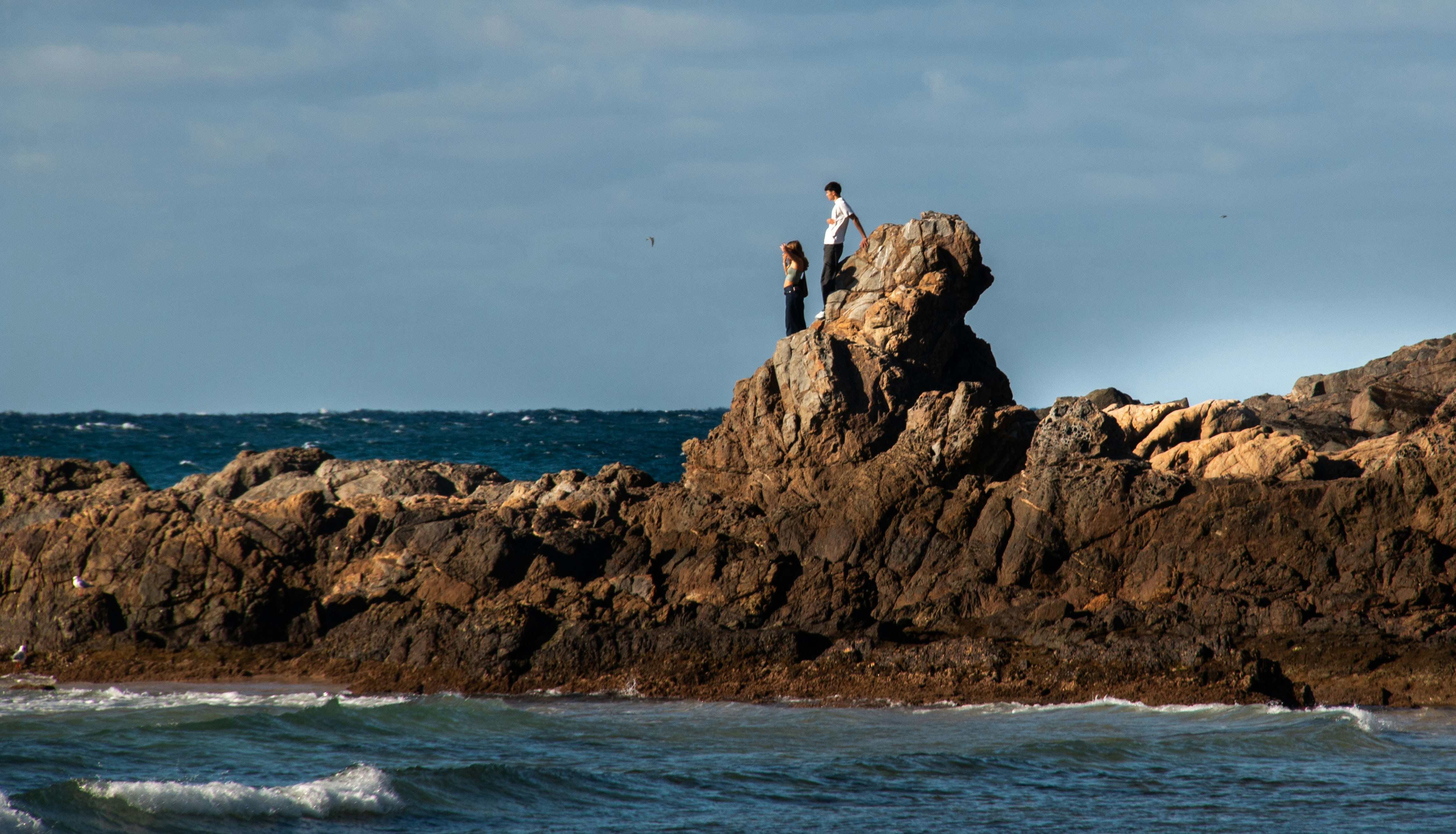 Two people stand on a rocky outcrop by the sea