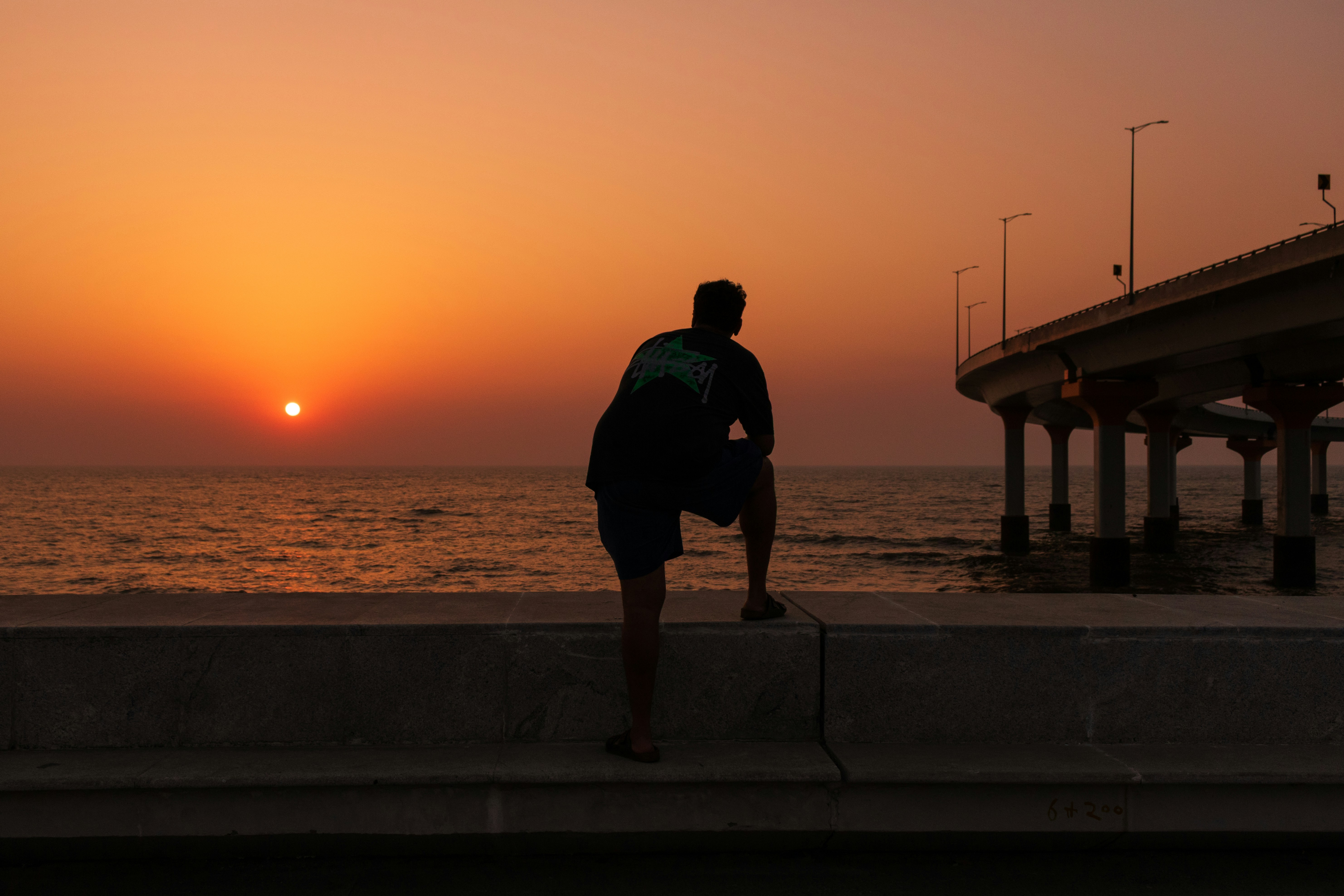 Man watches sunset over the ocean near a bridge.
