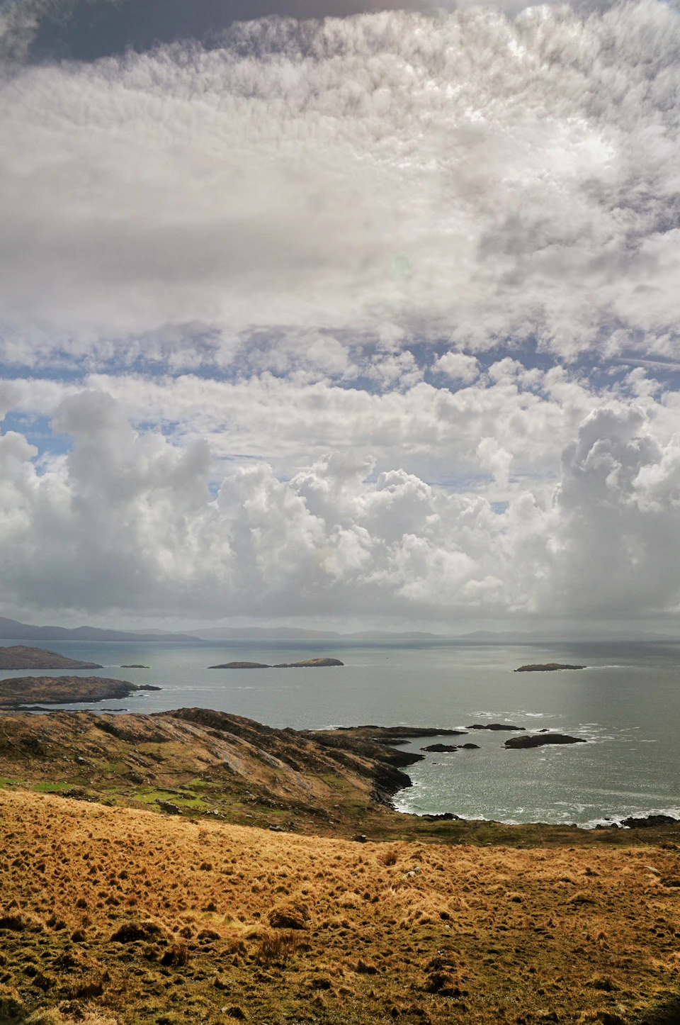 The image depicts a serene landscape with a panoramic view of a coastal area. The sky is filled with clouds, casting a soft light over the scene. Below, the ocean stretches out to meet the horizon at the lower right corner of the photo. The land features a mix of hills and valleys, creating a sense of depth in the image. The predominant colors are shades of green from the vegetation on the hills and browns from the earthy tones of the landscape.