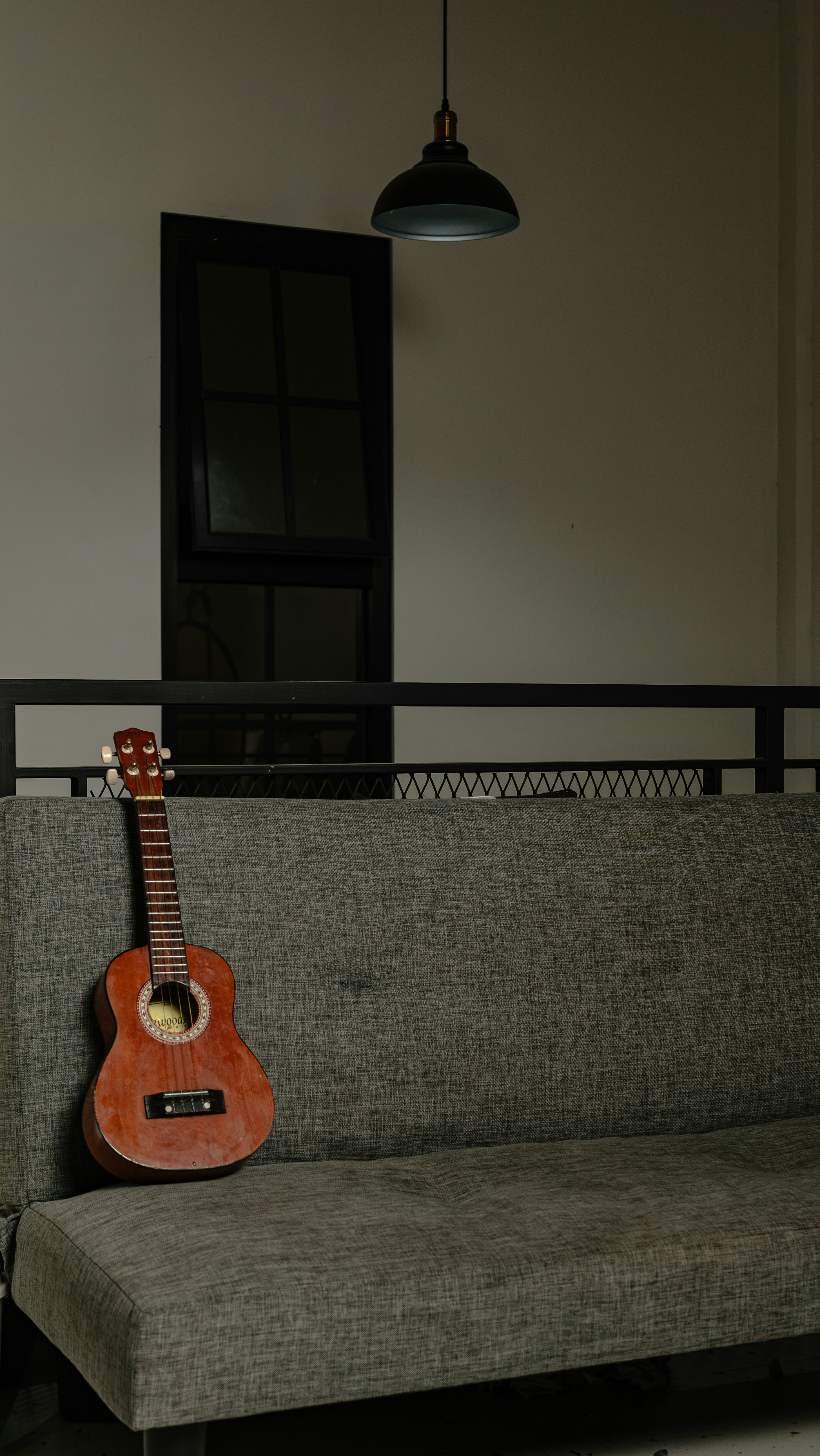 A ukulele rests on a grey sofa near a window.