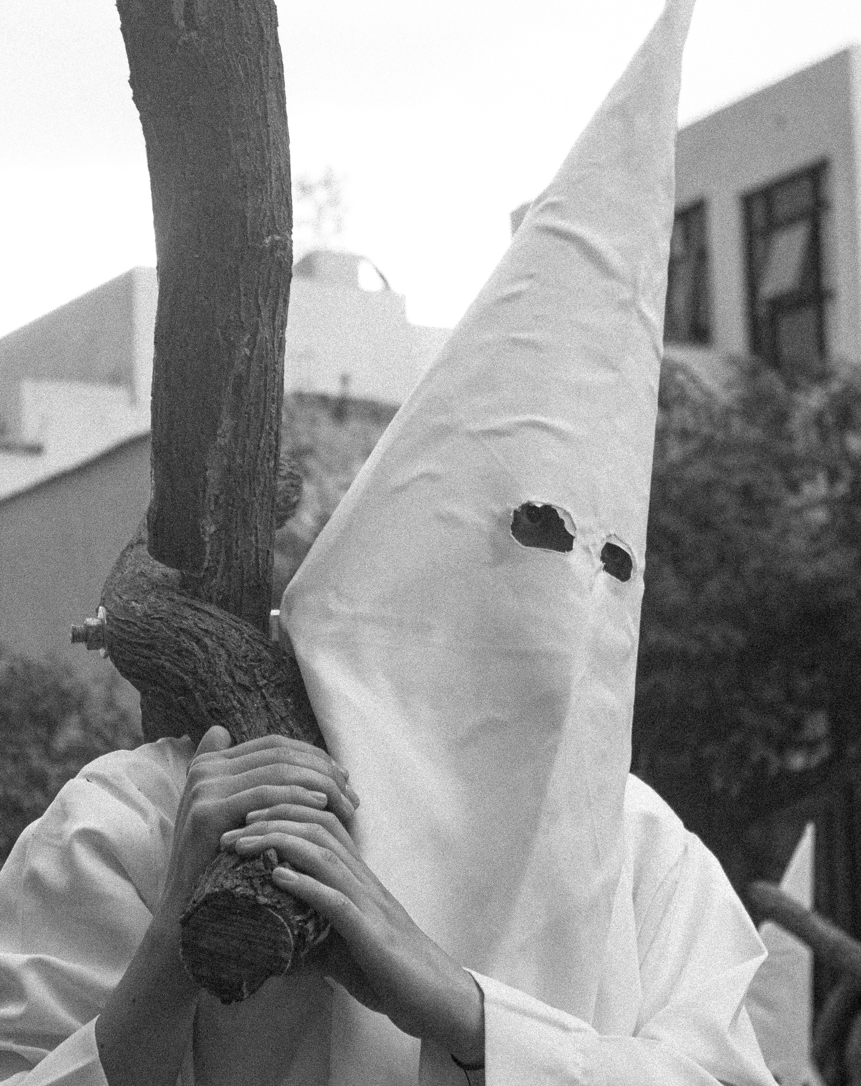 Person in a white hood carries a wooden cross.