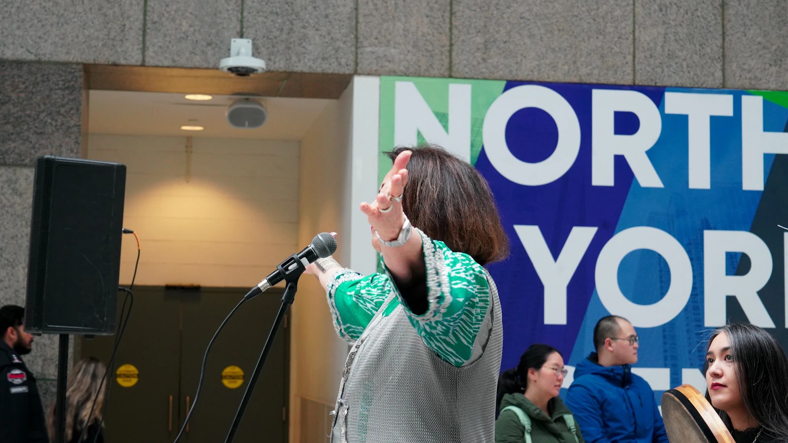 A woman speaking into a microphone in front of a gathered audience.