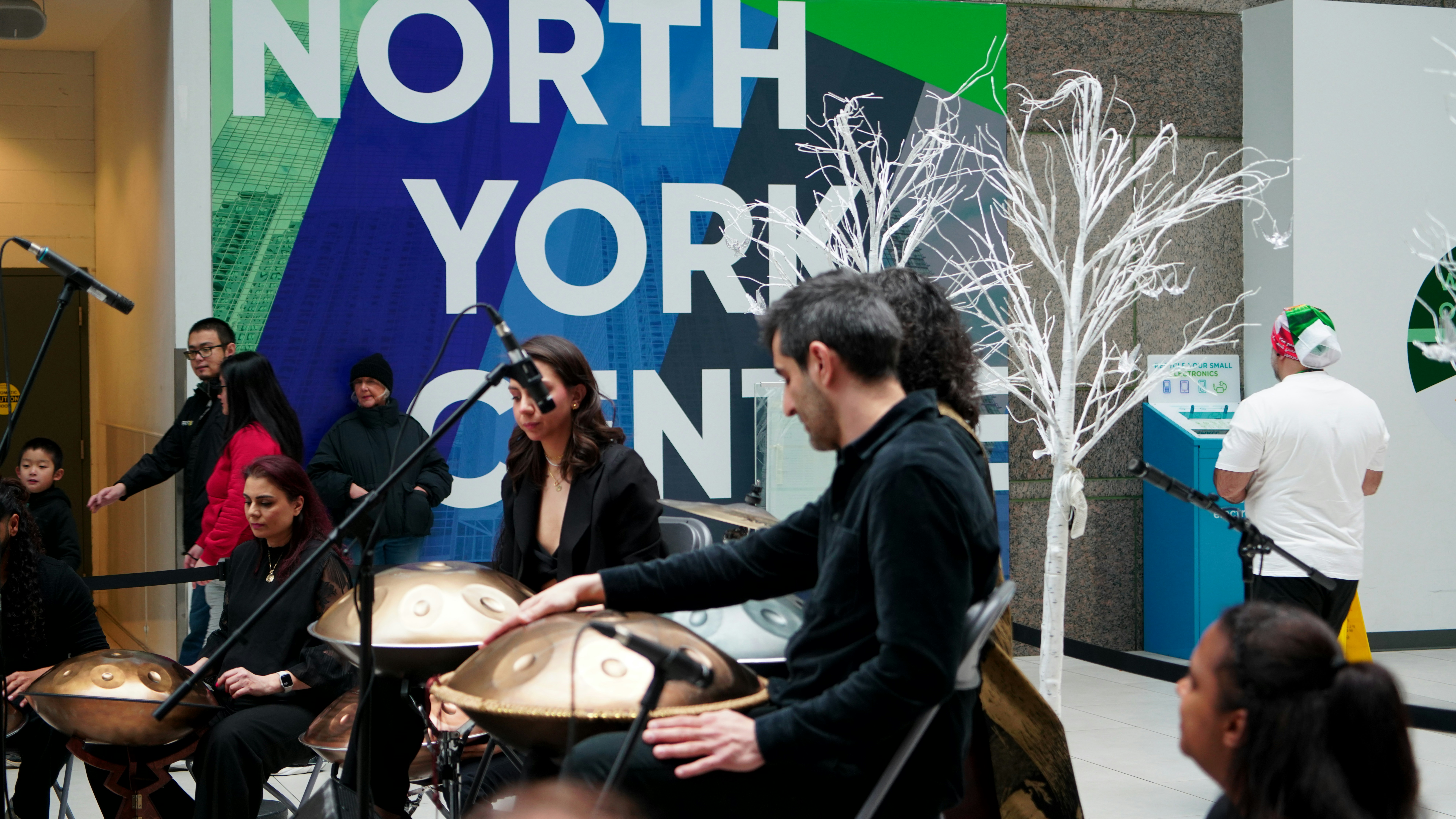 Musicians play handpan drums in front of a large sign.