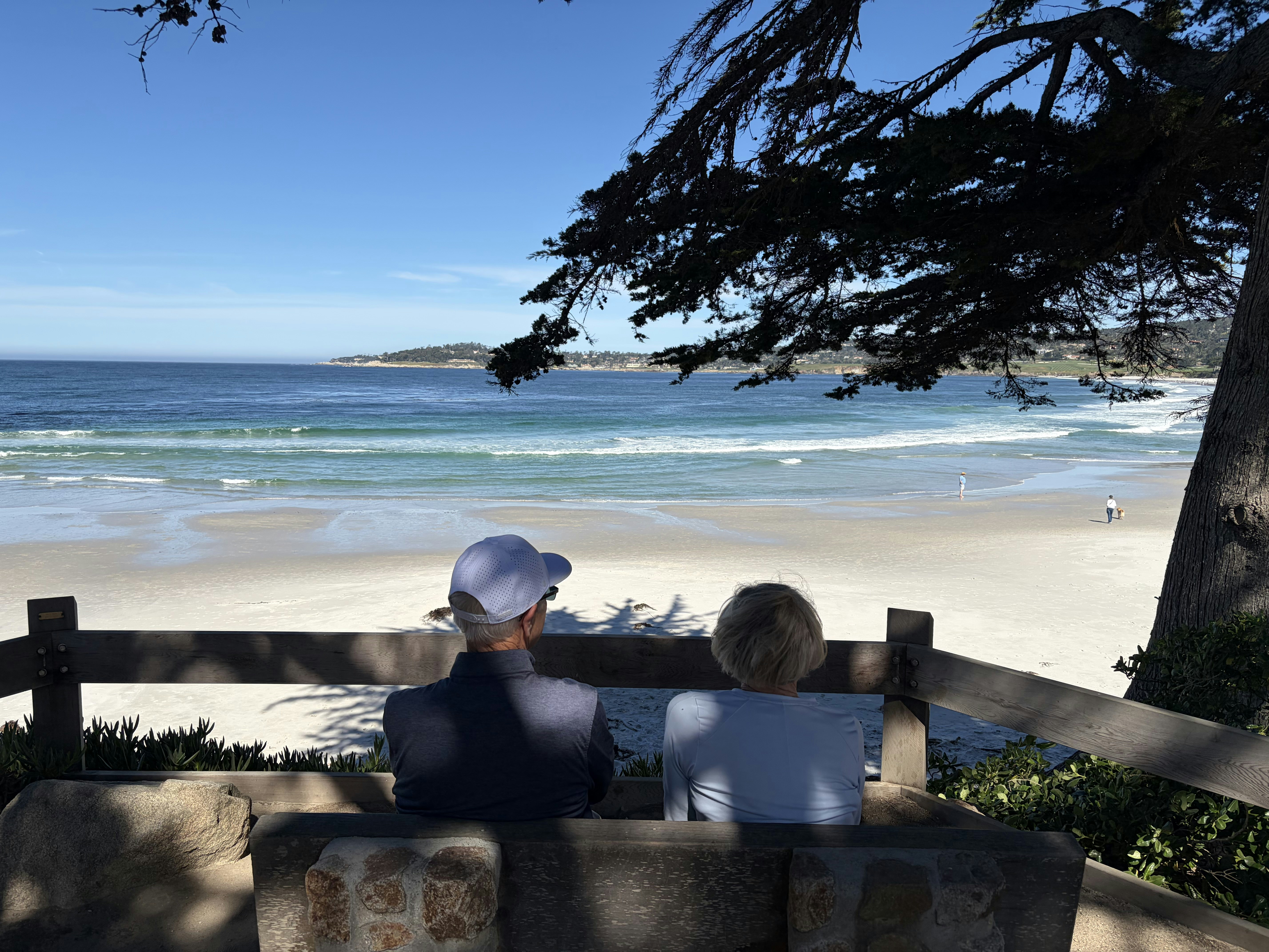 Zwei Personen sitzen auf einer Bank mit Blick auf einen Strand.