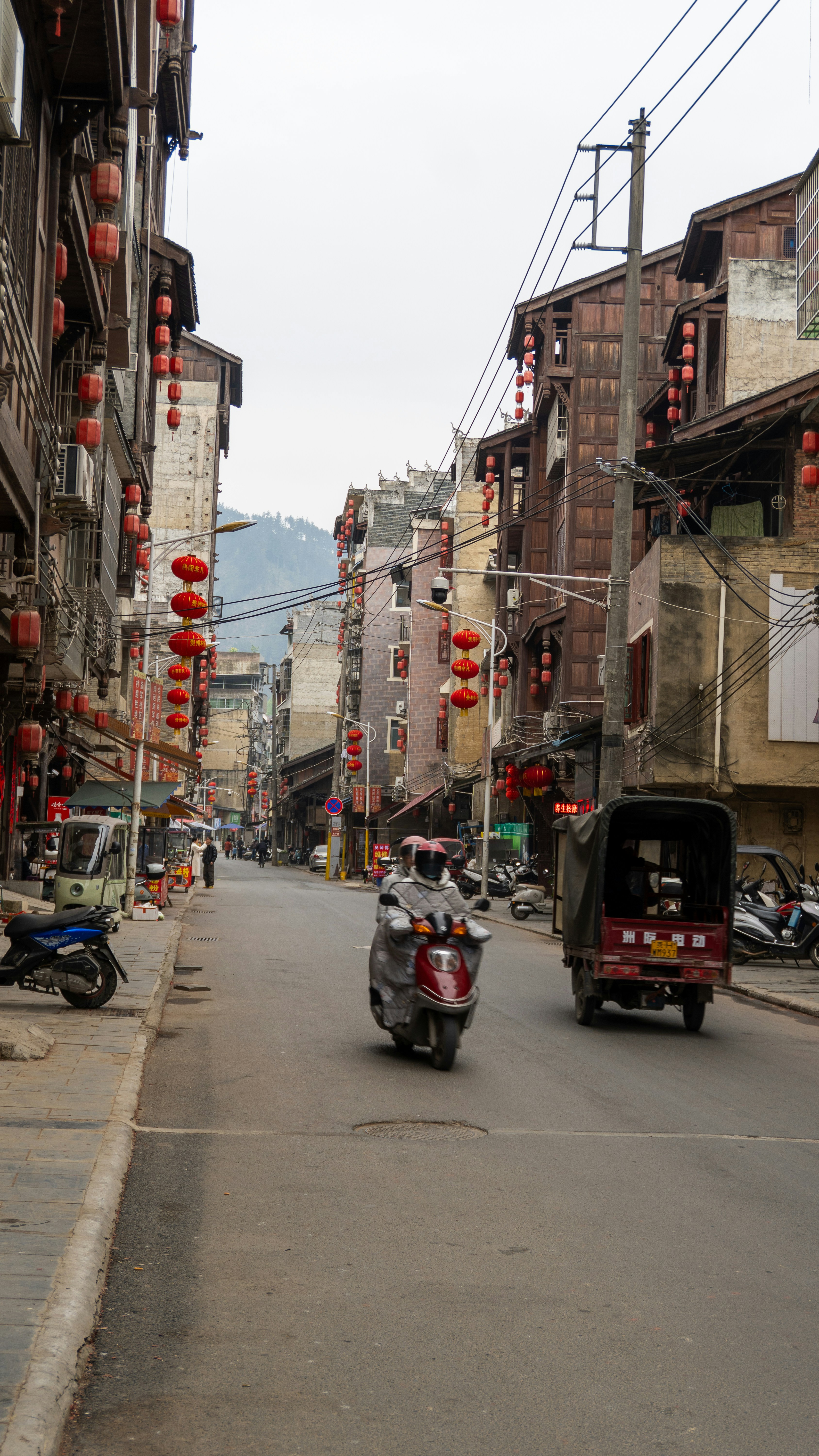 Le motociclette percorrono una strada fiancheggiata da edifici e lanterne.