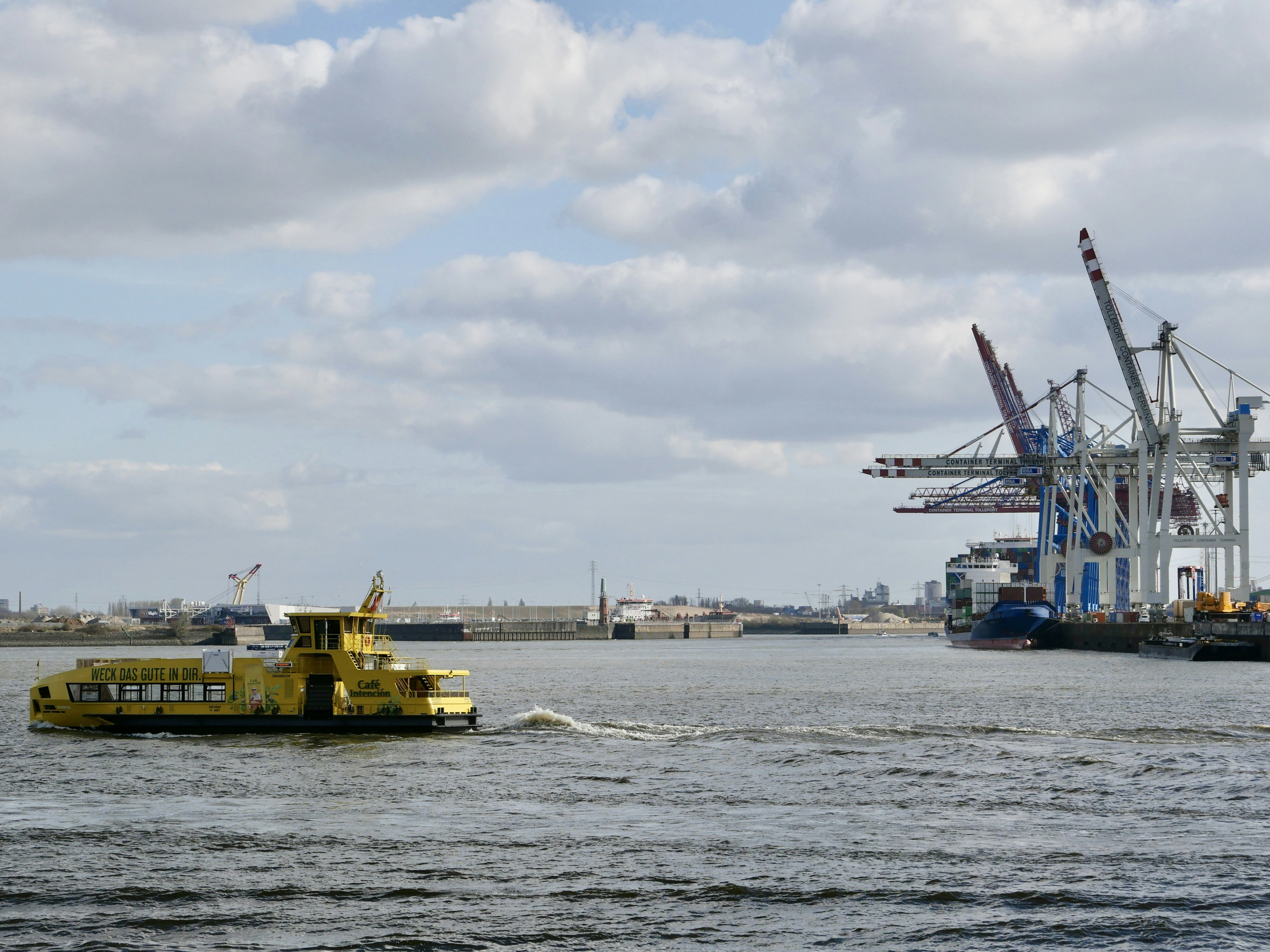 Yellow ferry boat sails on water near harbor cranes