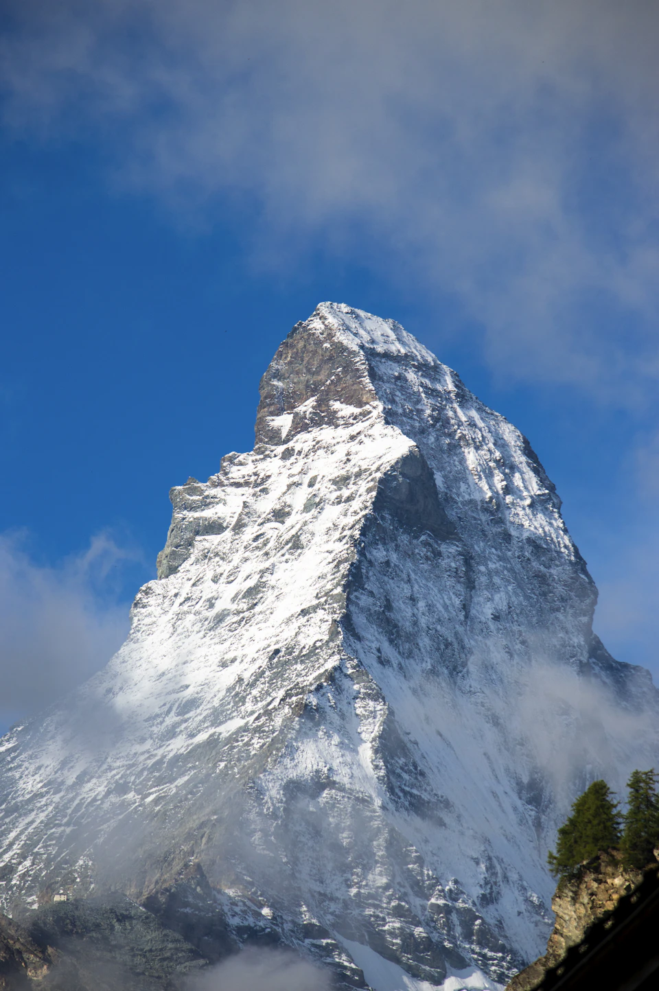 The image depicts a majestic snow-capped mountain peak against a clear blue sky with wispy clouds. The mountain, covered in a blanket of pristine white snow, stands tall and proud, its peak reaching towards the heavens. The perspective is from below, looking up at the mountain's grandeur, emphasizing its towering presence.

In the distance, there are trees dotting the landscape, adding depth to the scene. The image does not contain any discernible text or countable objects, making it a captivating snapshot of nature's beauty and power.