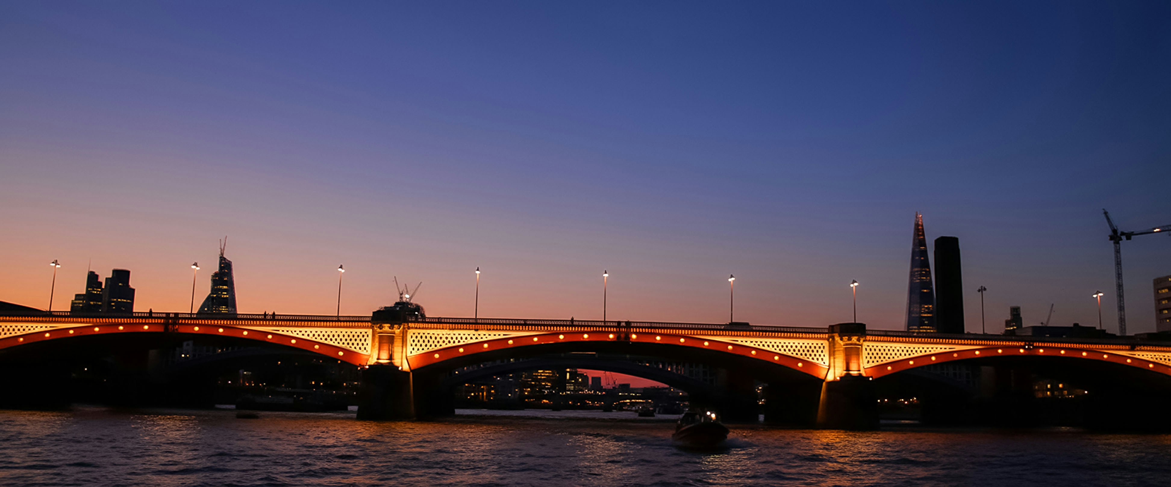 Illuminated bridge over river at dusk with city skyline.