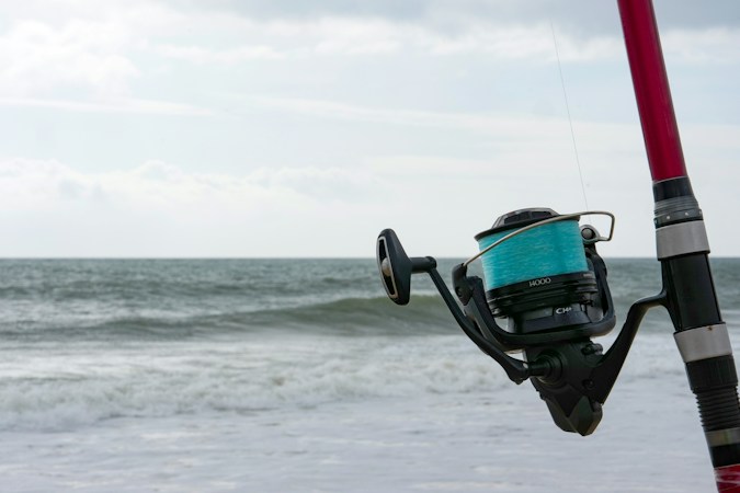 Spinning reel spooled with bright teal line on a red surf rod, gray-green waves rolling onto a beach behind it