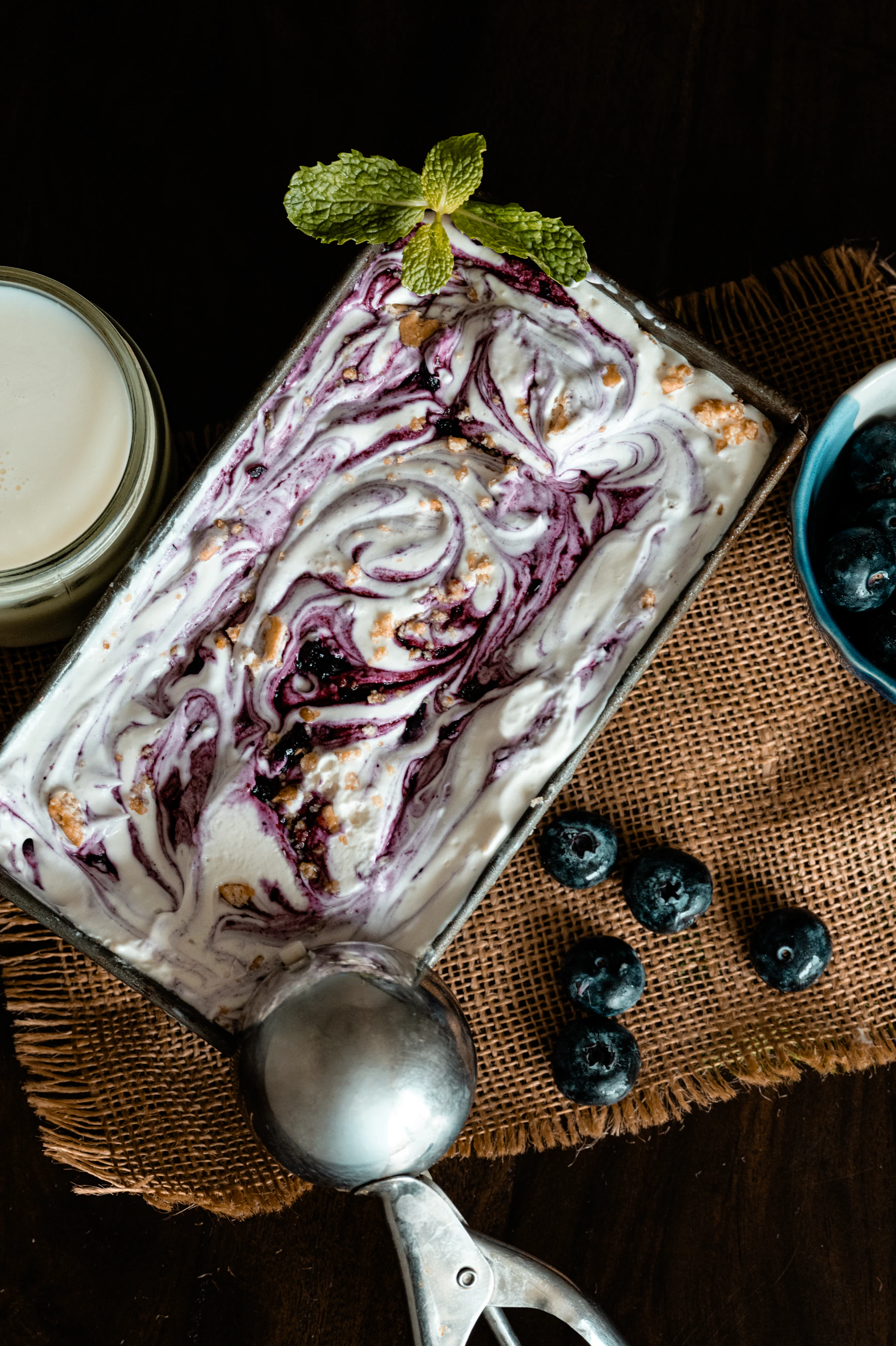 A loaf pan of white ice cream swirled with blueberry, a metal scoop and fresh blueberries on burlap
