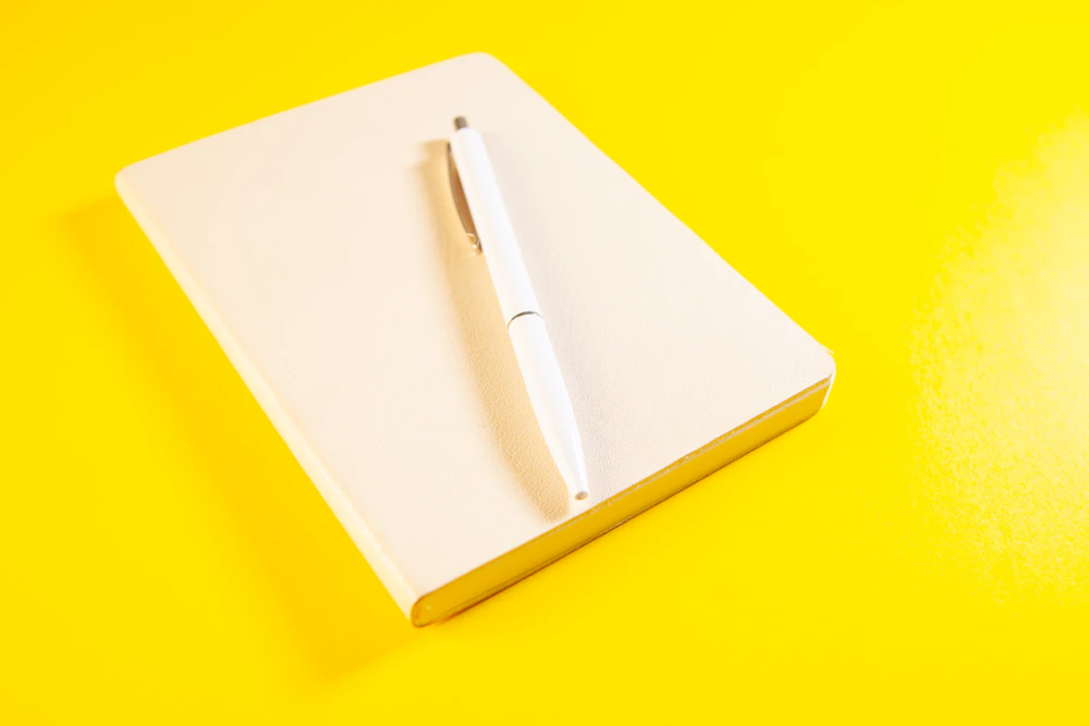 Ein weißer Stift liegt auf einem geschlossenen Notizbuch vor gelbem Hintergrund