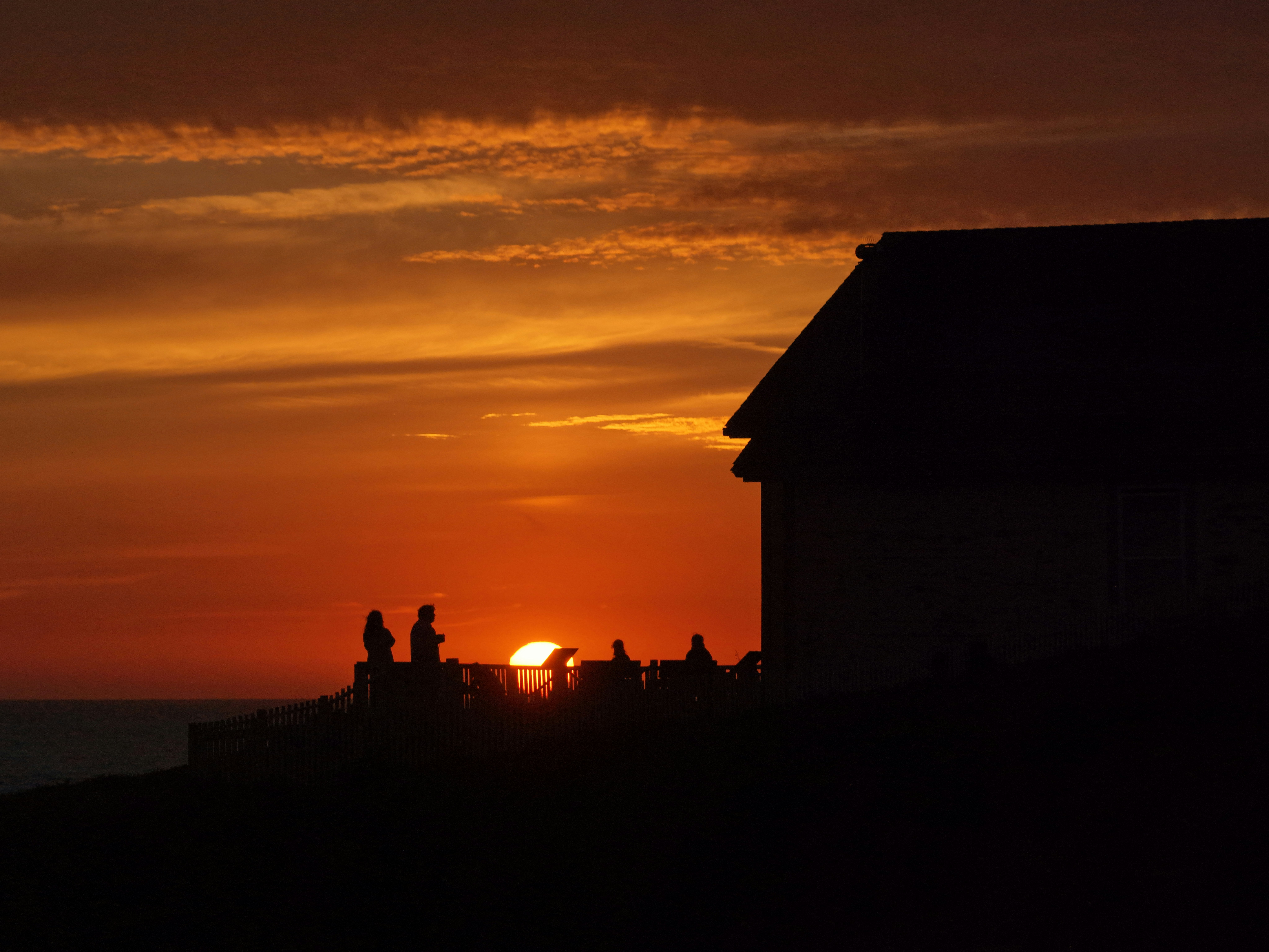 Figuras en silueta observan la puesta de sol junto a un edificio.