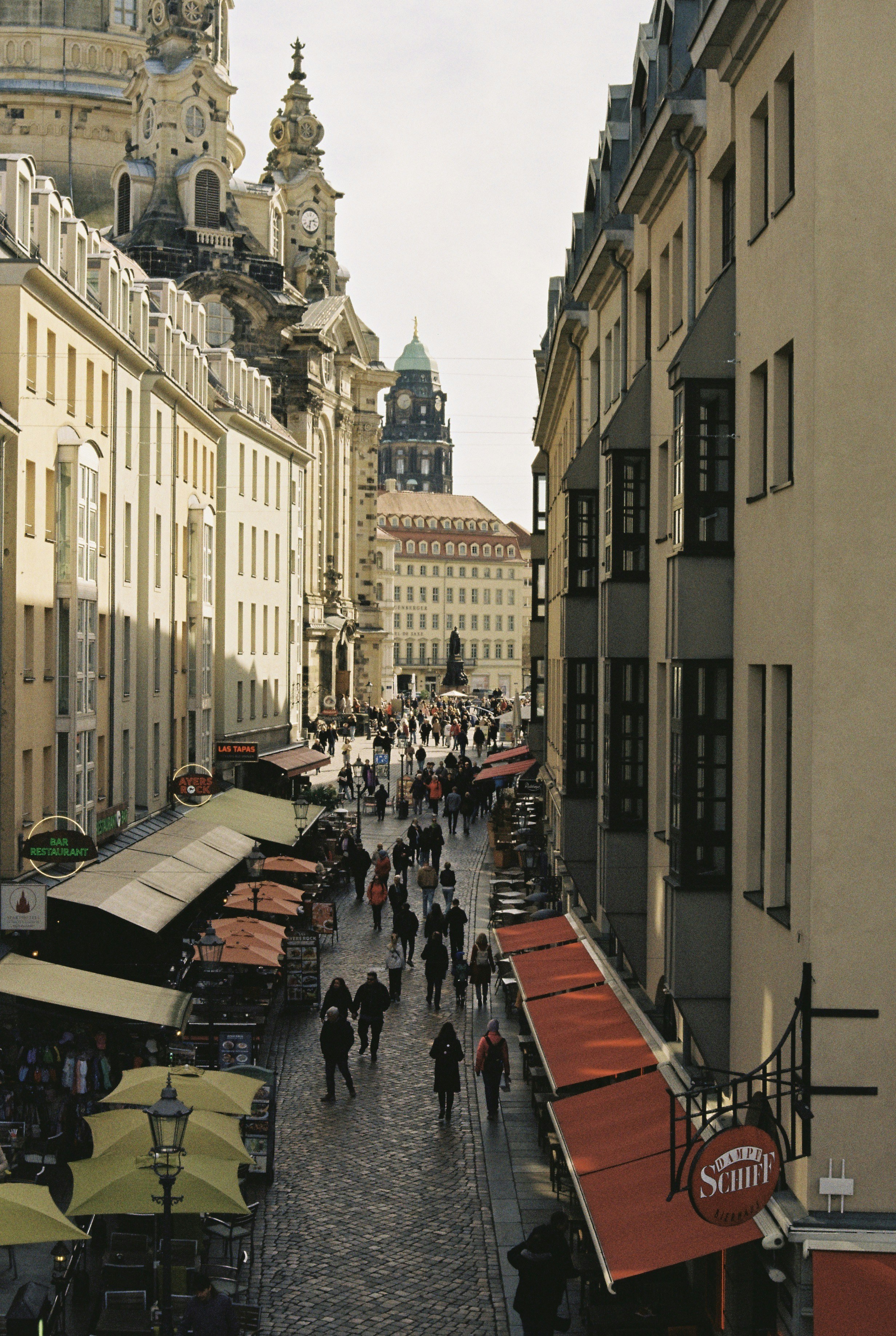 Calle empedrada llena de gente, bordeada de tiendas y cafeterías.