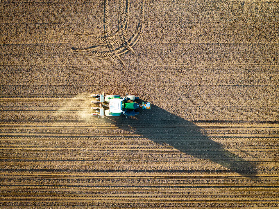 Tractor plows a large field with kicking up dust in an aerial view