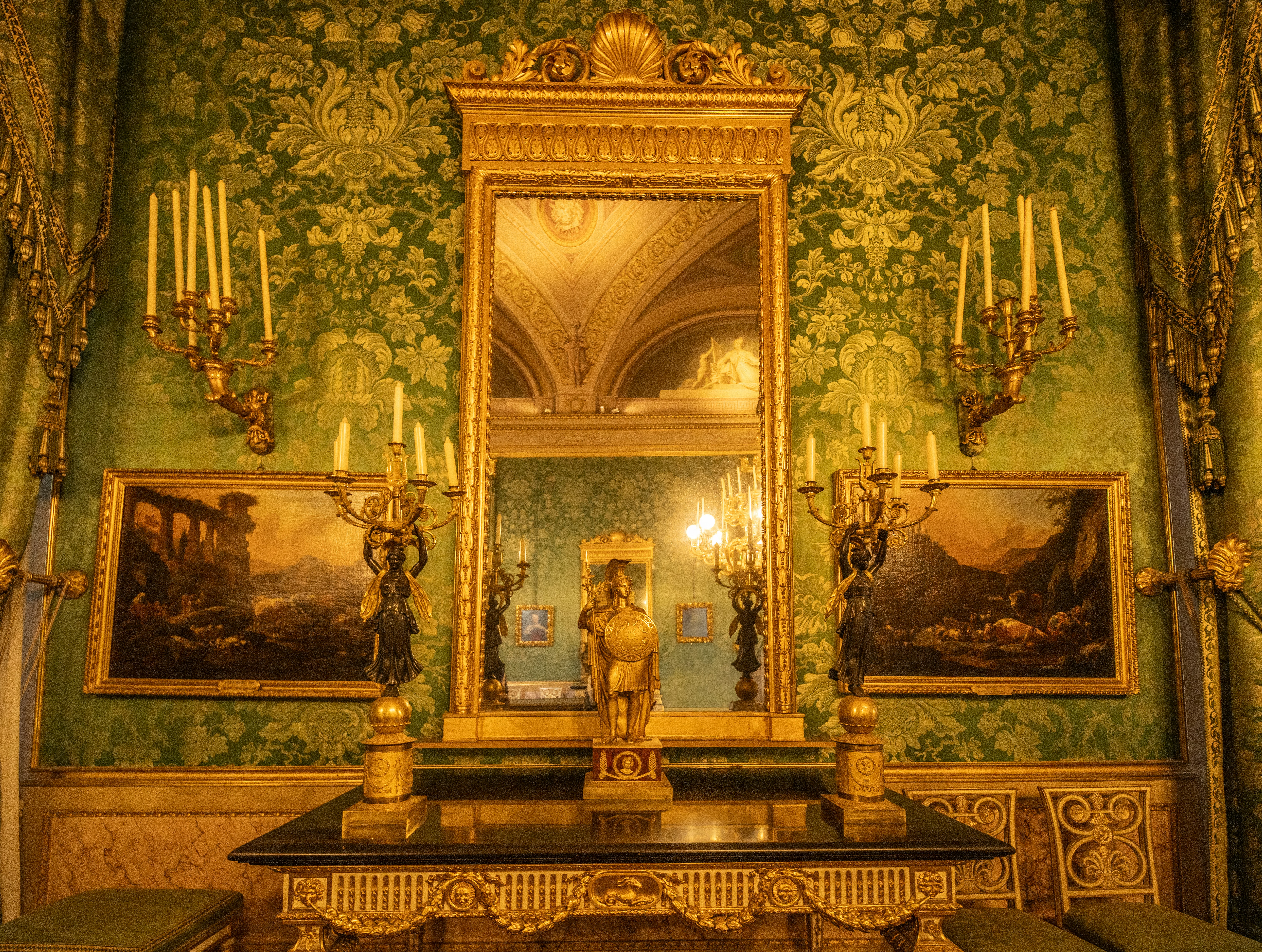 Ornate golden mirror reflecting a grand hall with paintings.