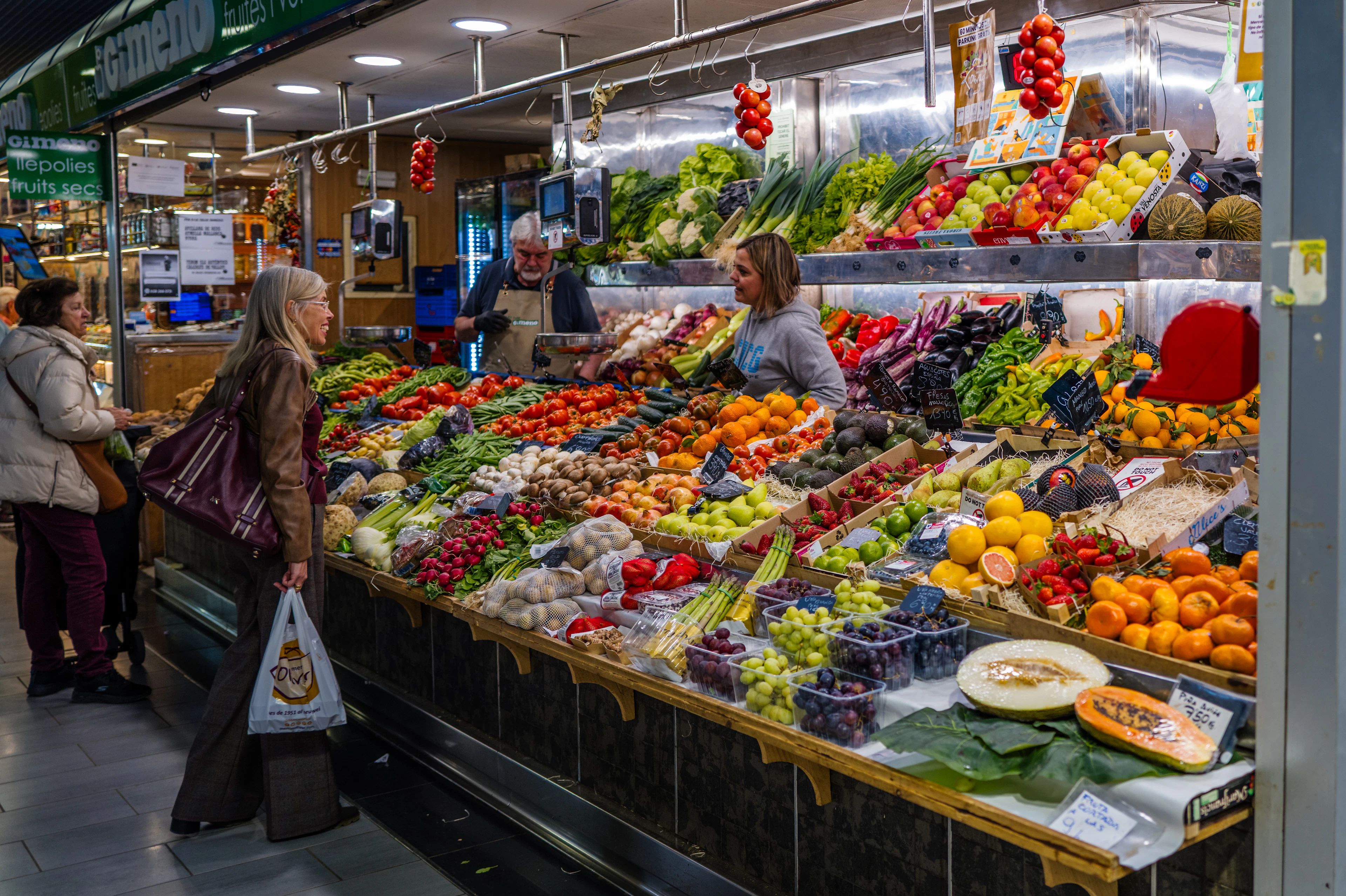 A produce stall inside a covered market hall in central Palma de Mallorca, its counter banked with tomatoes, peppers, citrus, grapes and cut melon while a stallholder in an apron serves a customer