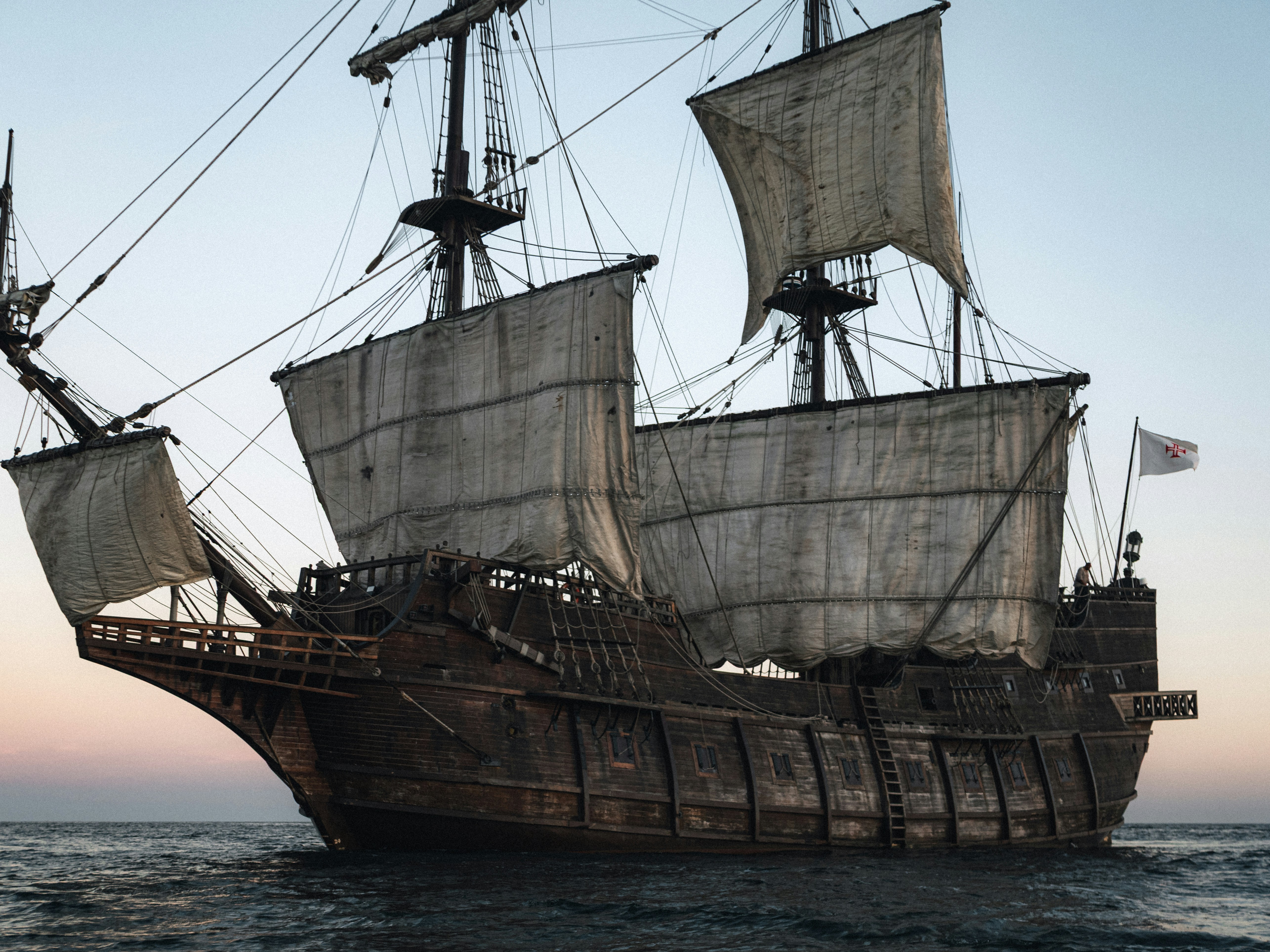 Historic wooden sailing ship at sea during sunset, with calm water, soft light and a dramatic silhouette against the horizon.