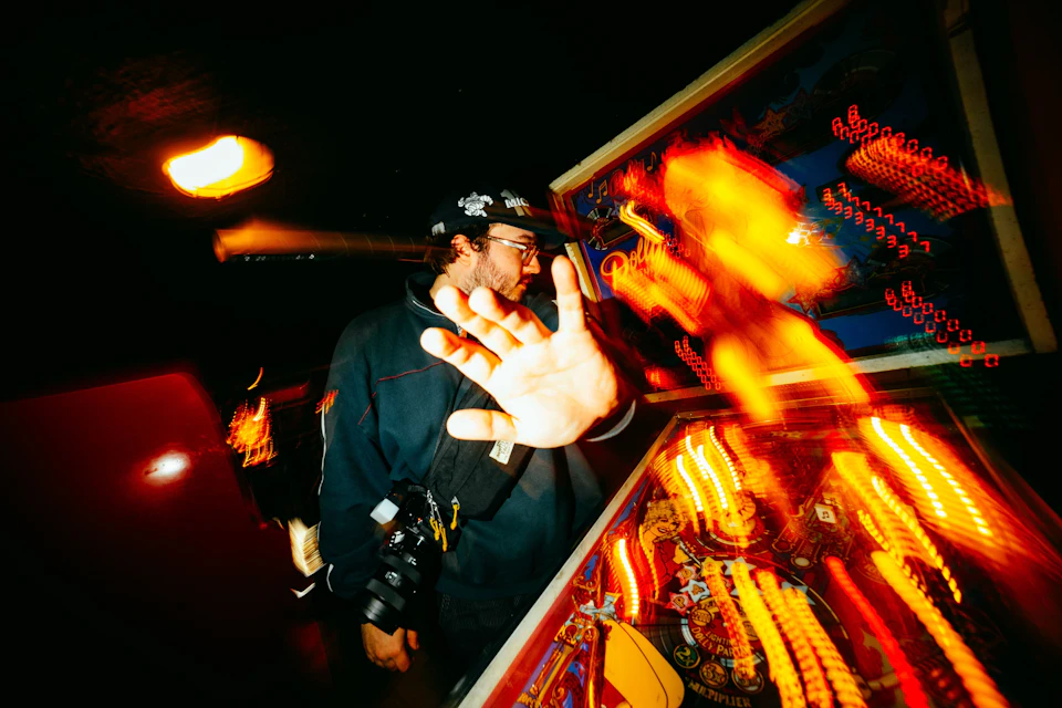 The image depicts a man standing in front of a pinball machine, with his hand raised to hit the ball. The pinball machine is illuminated by bright lights and features a vibrant red background that contrasts with the black and white elements within it. A blurred figure can be seen on the left side of the frame, adding depth to the scene.