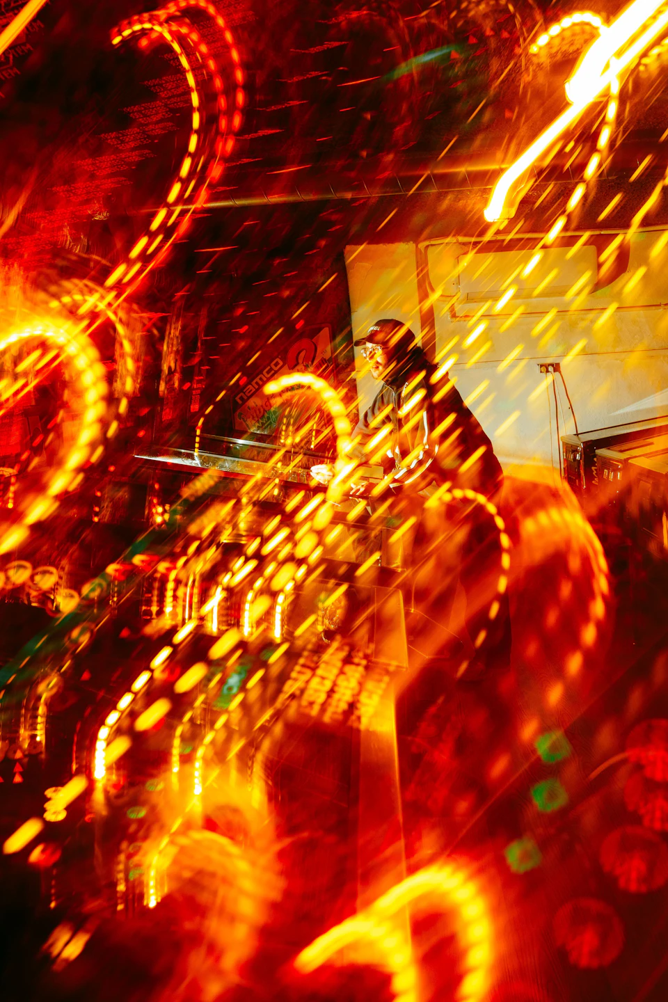 The image depicts a vibrant scene at night, with a building illuminated by bright orange and yellow lights that create an ethereal glow. The perspective is from below, looking up towards the building's facade, which adds to the sense of grandeur and scale. In the foreground, there are blurred figures of people, suggesting movement or activity within the scene.