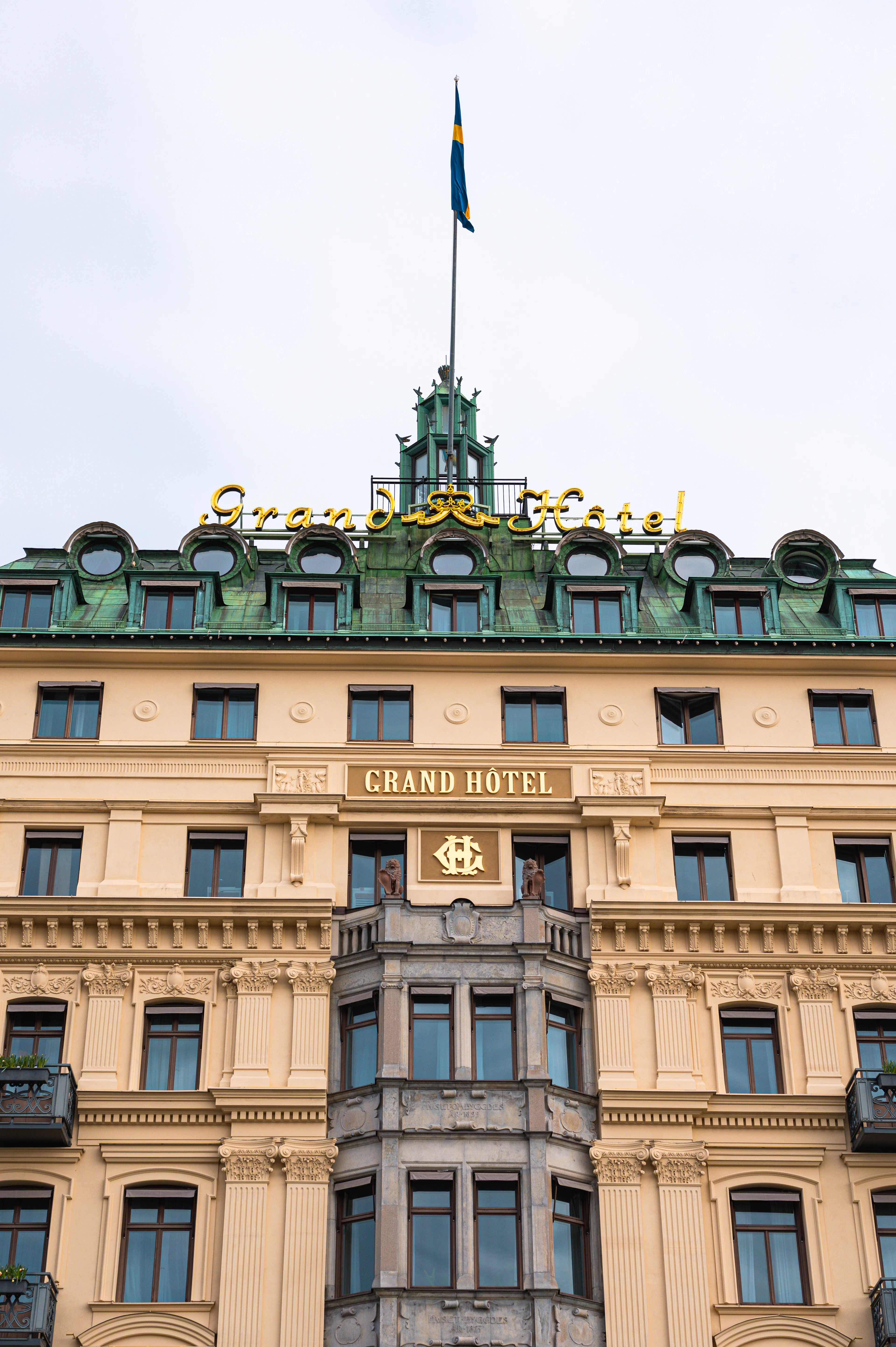 The cream stone facade of the Grand Hôtel in Stockholm seen from below, its gold neon script and verdigris copper roof crowned by a Swedish flag.
