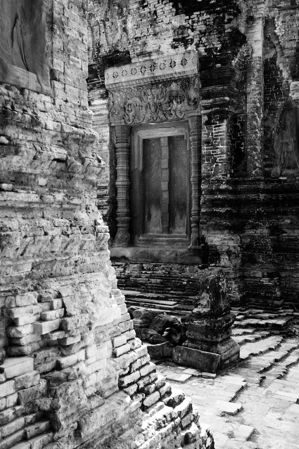 The image depicts a temple nestled within an ancient stone wall, showcasing intricate carvings and designs that hint at its historical significance. The perspective is from below, looking up towards the temple's towering presence against the sky. A small window punctuates one of the walls, adding to the architectural details. The black and white color scheme enhances the timeless quality of the scene, emphasizing the age-old structures and their enduring beauty.