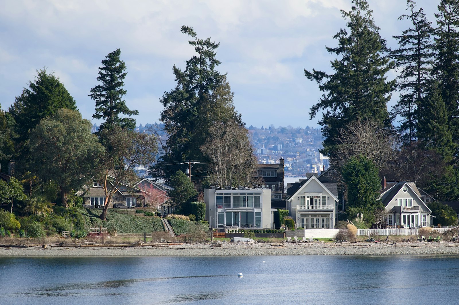 Waterfront homes among tall Douglas firs across a saltwater channel, a city hillside rising behind