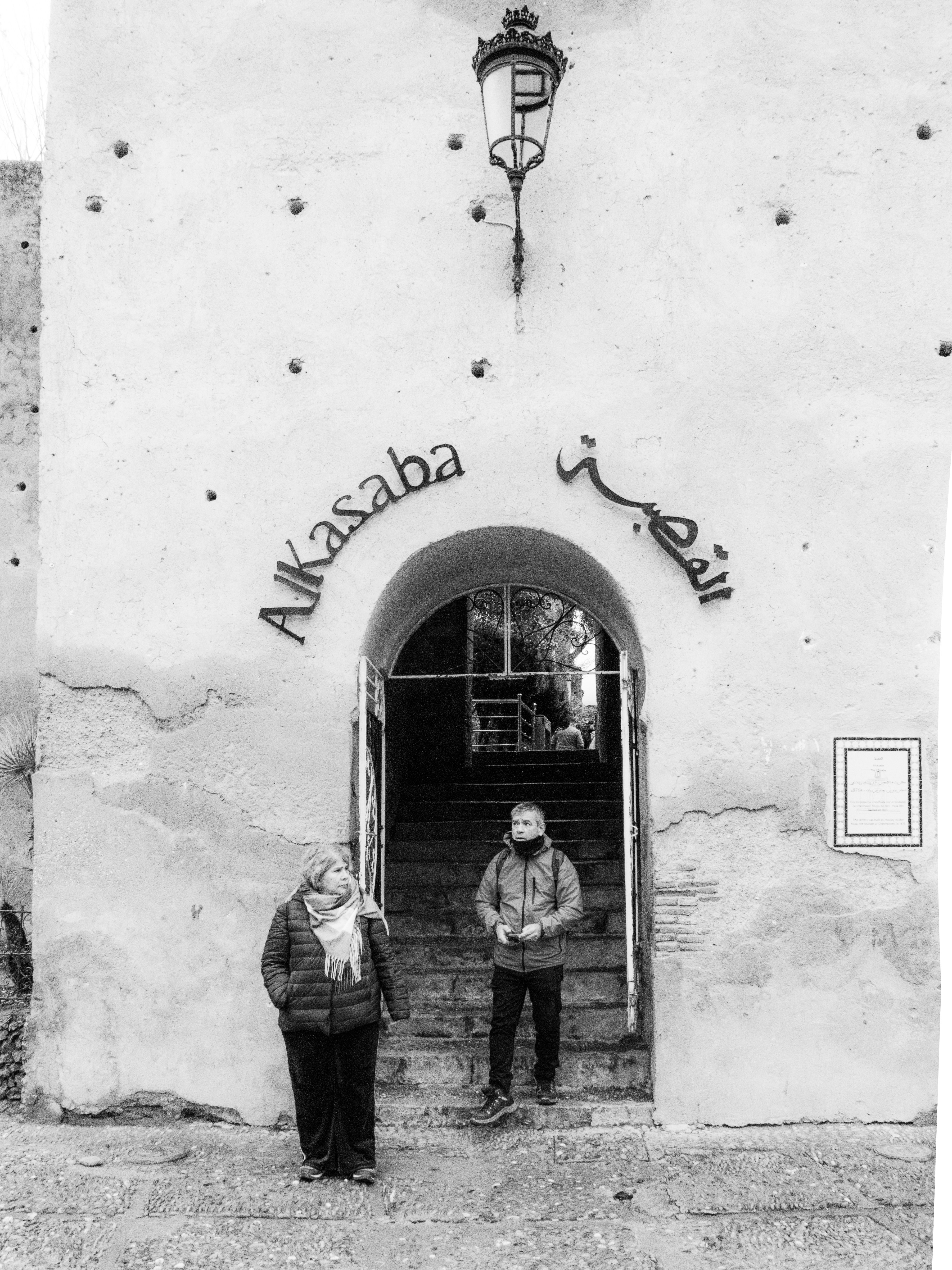 Two people stand by an arched doorway with arabic inscription.