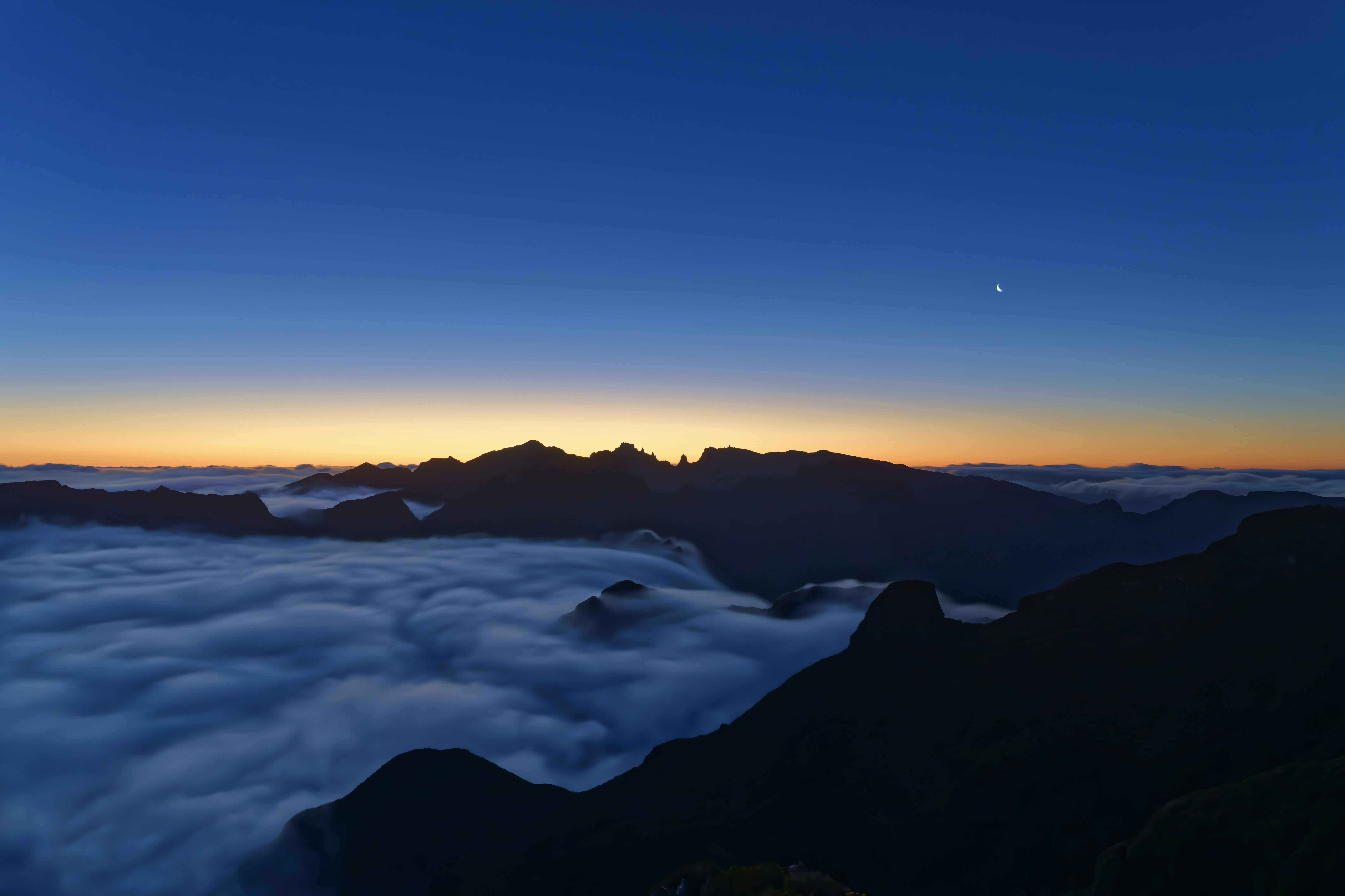 Mountains above clouds at dusk with a sliver of moon.
