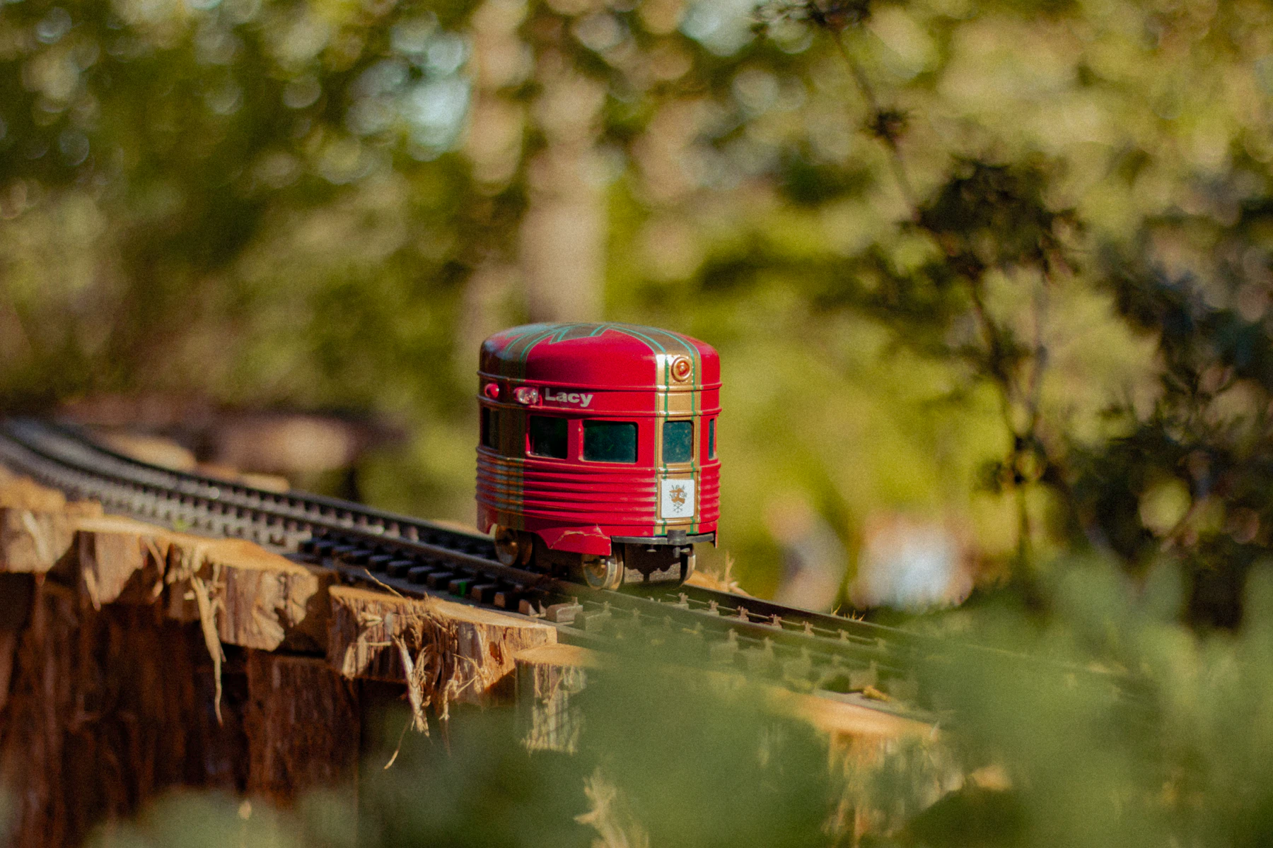 A red model passenger car on a curve of raised track, with sunlit trees blurred behind