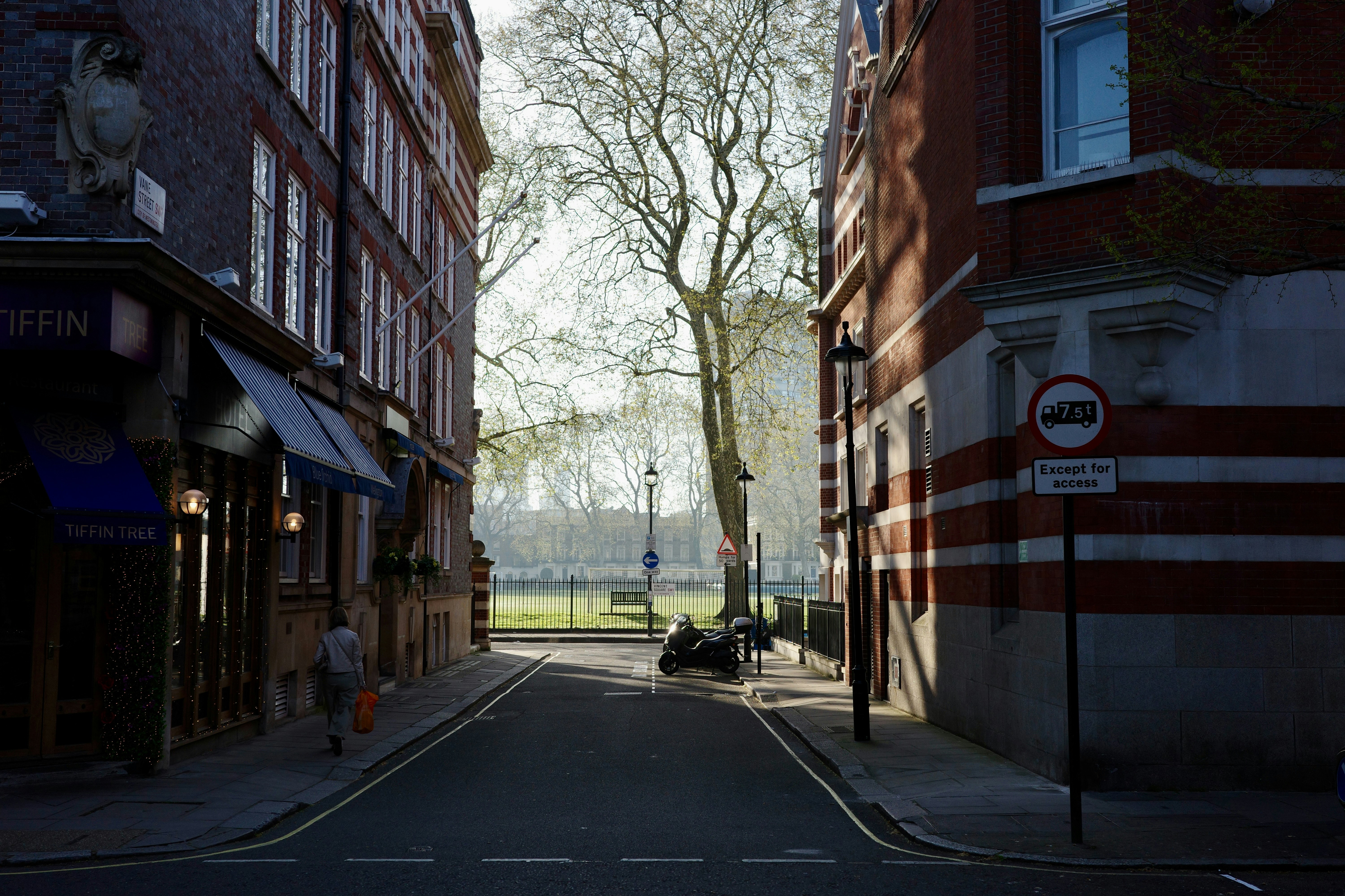 A narrow street between brick buildings with a park beyond