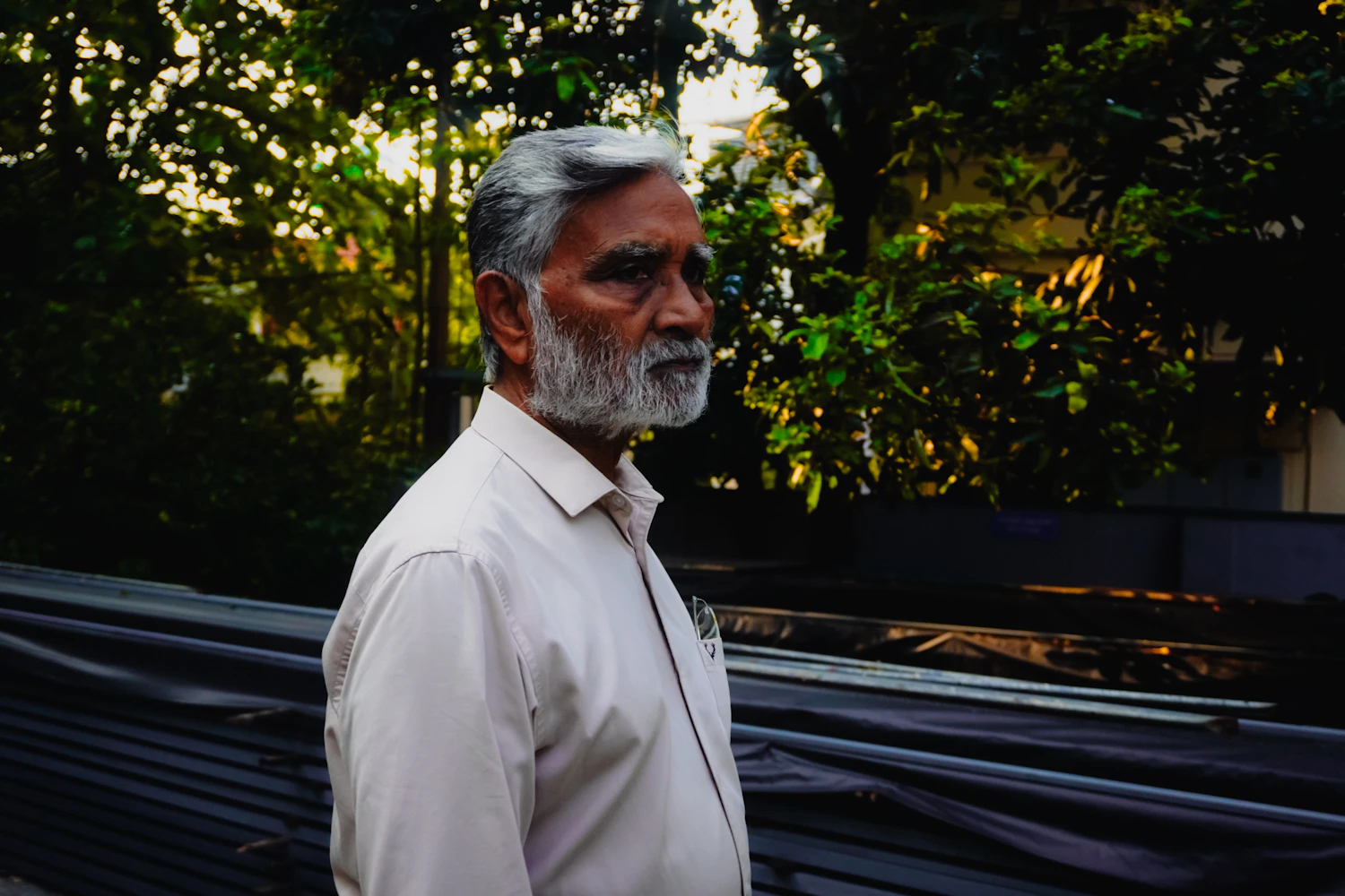 A man with a white beard in a pale shirt, half turned away, standing in front of stacked sheet material