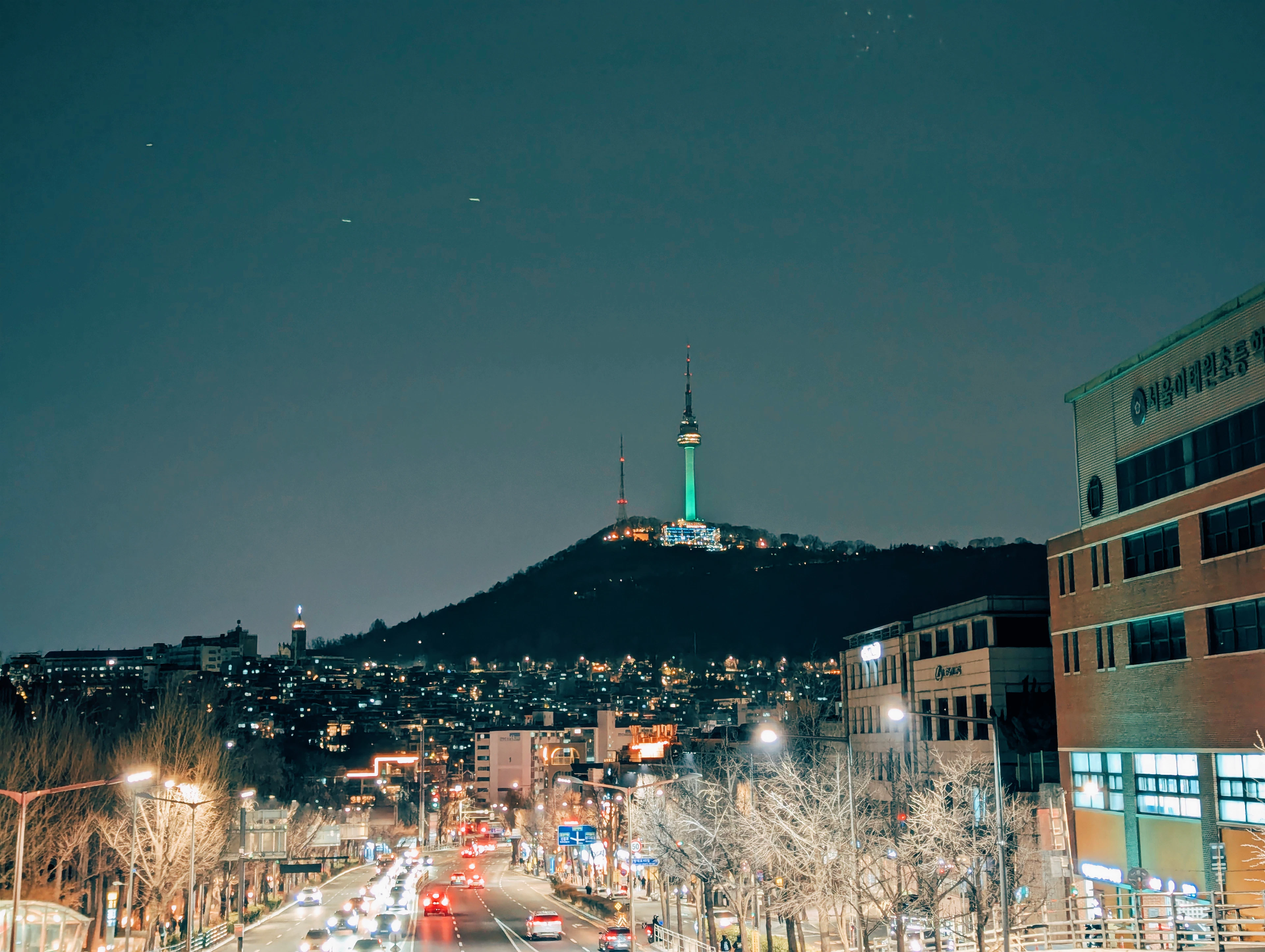 N Seoul Tower lit up on Namsan mountain after dark, with the illuminated streets and apartment blocks of Seoul spread out below.